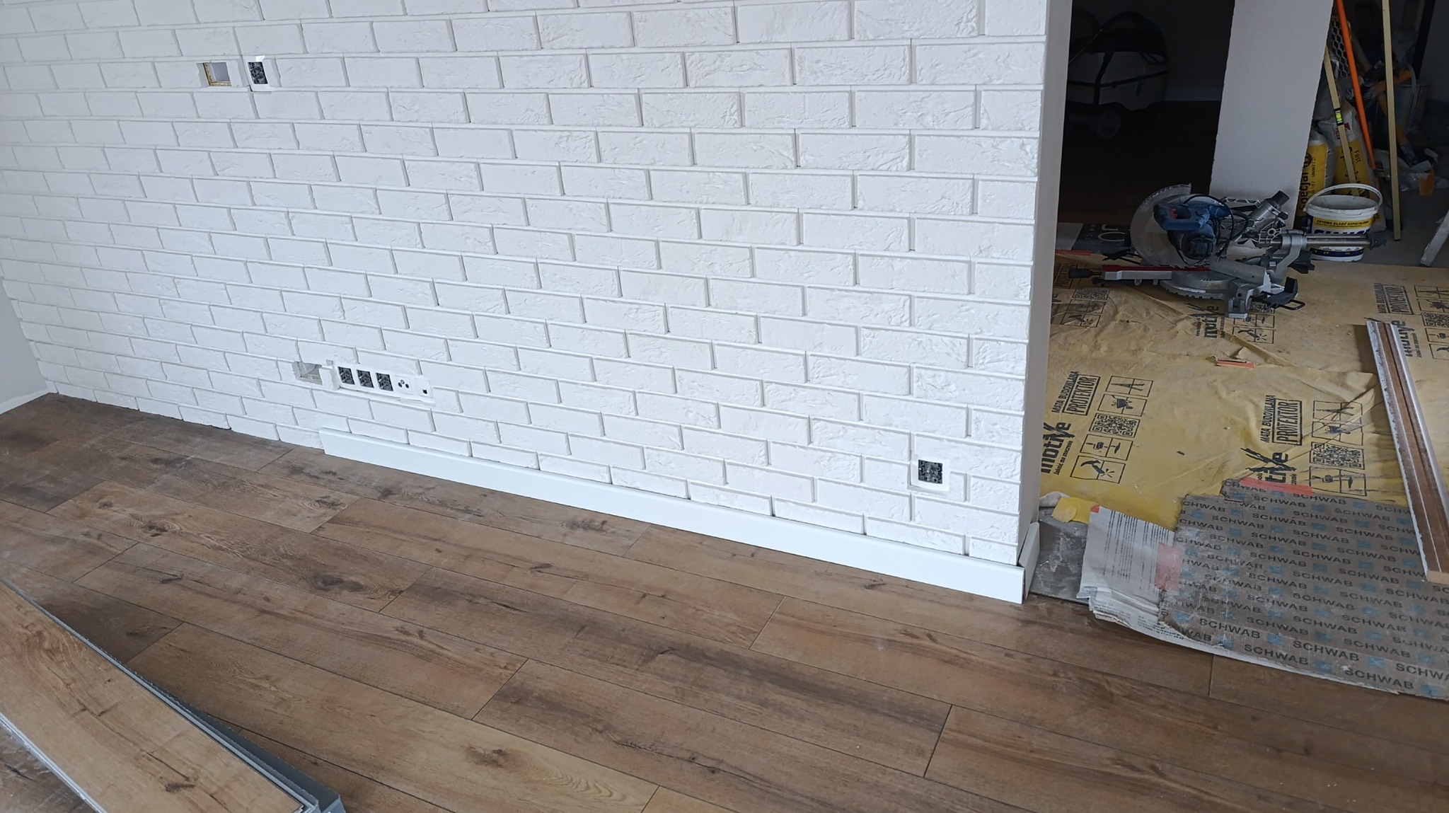 Biała ściana z cegły dekoracyjnej, brązowa podłoga z paneli, widoczne puszki elektryczne i listwa przypodłogowa. W tle narzędzia na podłodze zabezpieczonej folią.