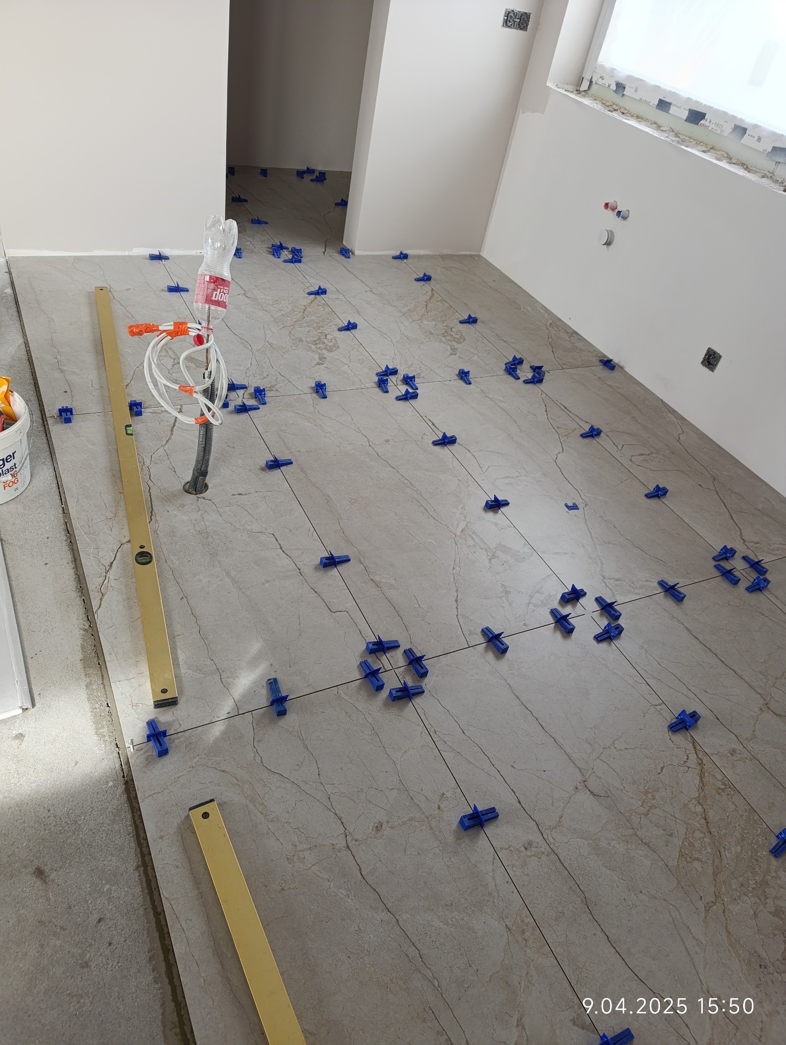 Układanie beżowych płytek podłogowych z użyciem niebieskich klipsów poziomujących. Widoczna poziomica i rurka wystająca z podłogi. Prace w trakcie realizacji.