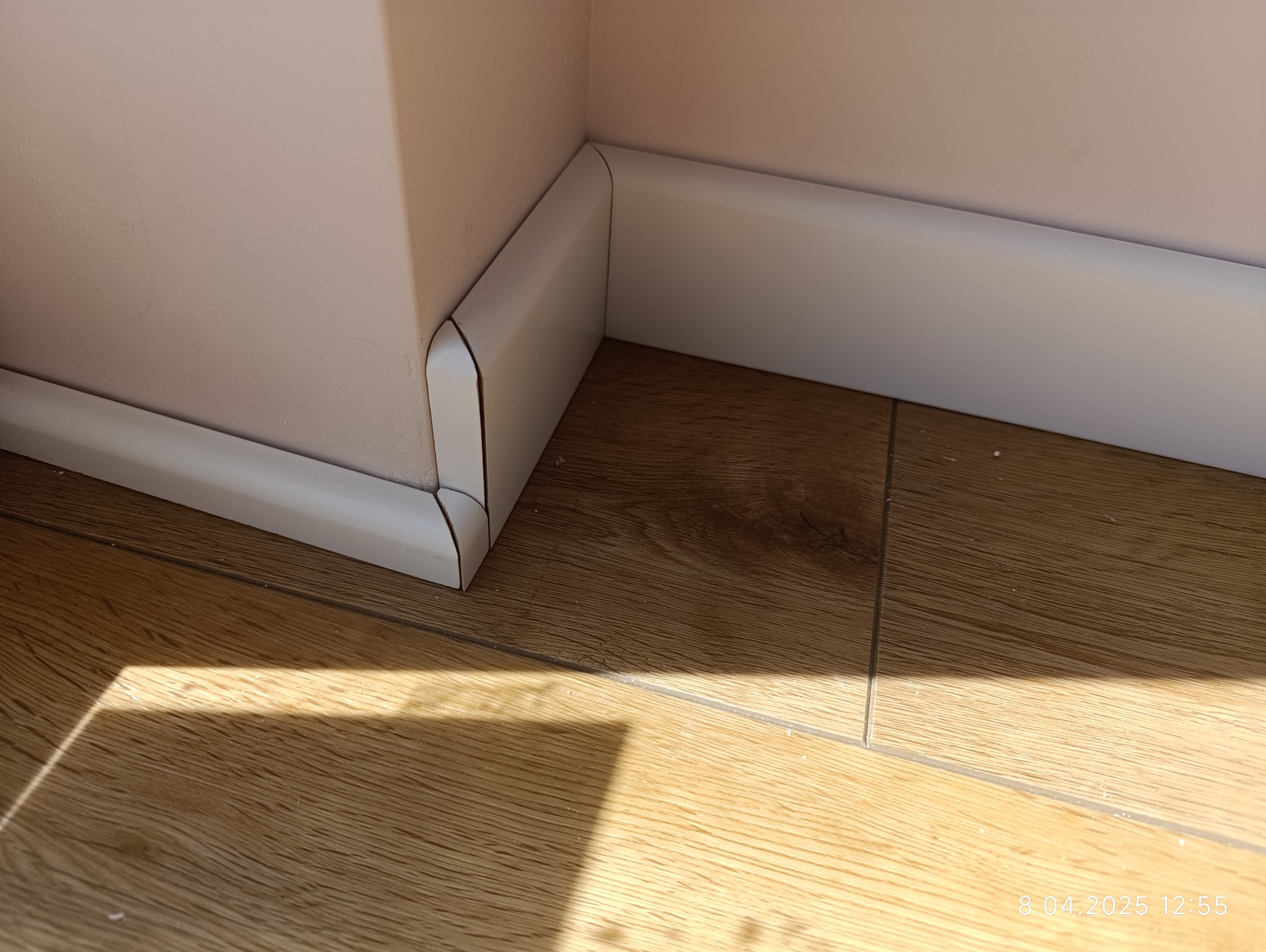Białe listwy przypodłogowe zamontowane w rogu pomieszczenia, na drewnopodobnej podłodze. Widoczne połączenie listew i fragment ściany w jasnym odcieniu.