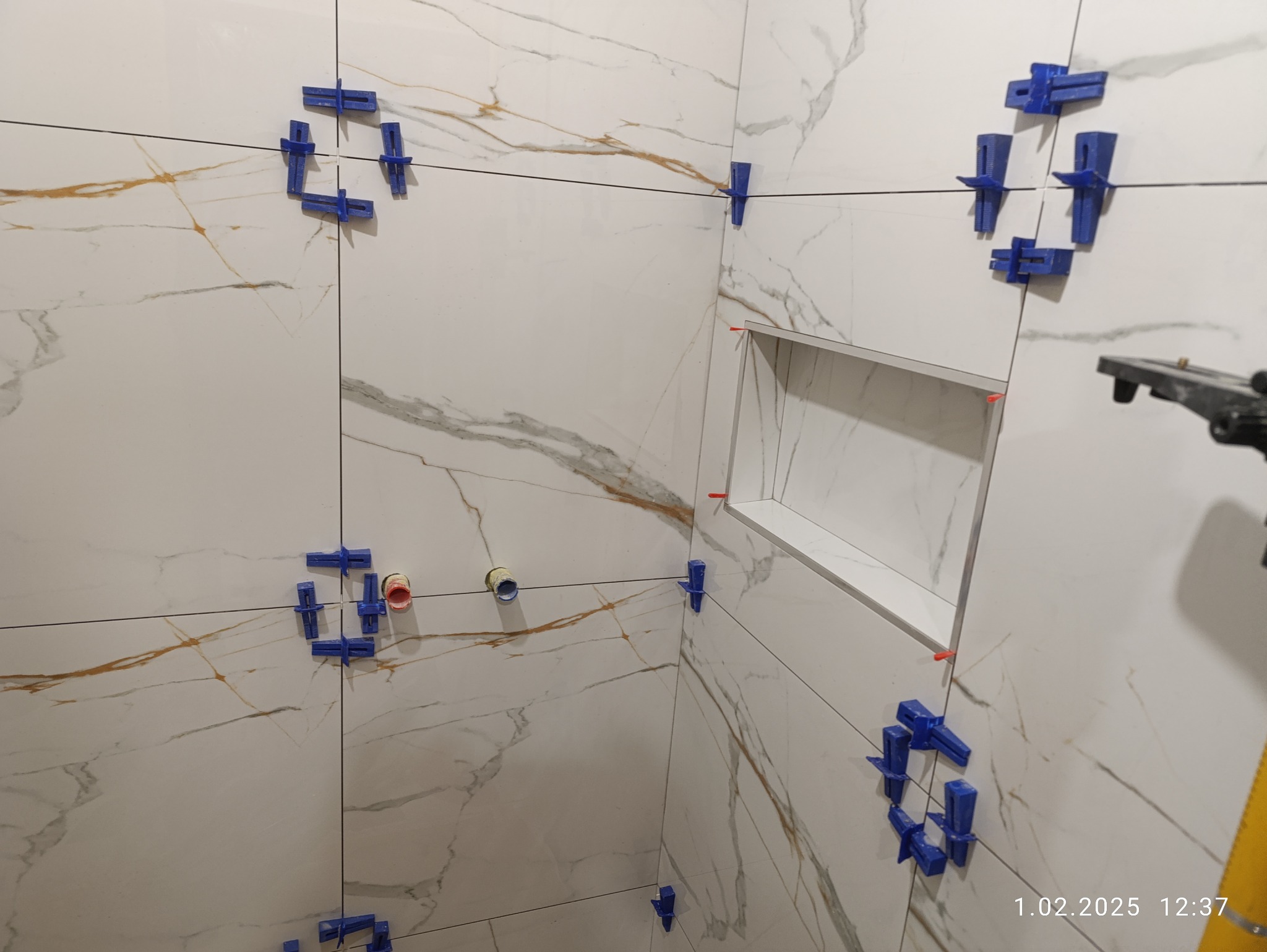 Białe płytki imitujące marmur z niebieskimi klipsami poziomującymi, widoczna wnęka w ścianie i fragment poziomicy. Etap prac wykończeniowych w łazience.