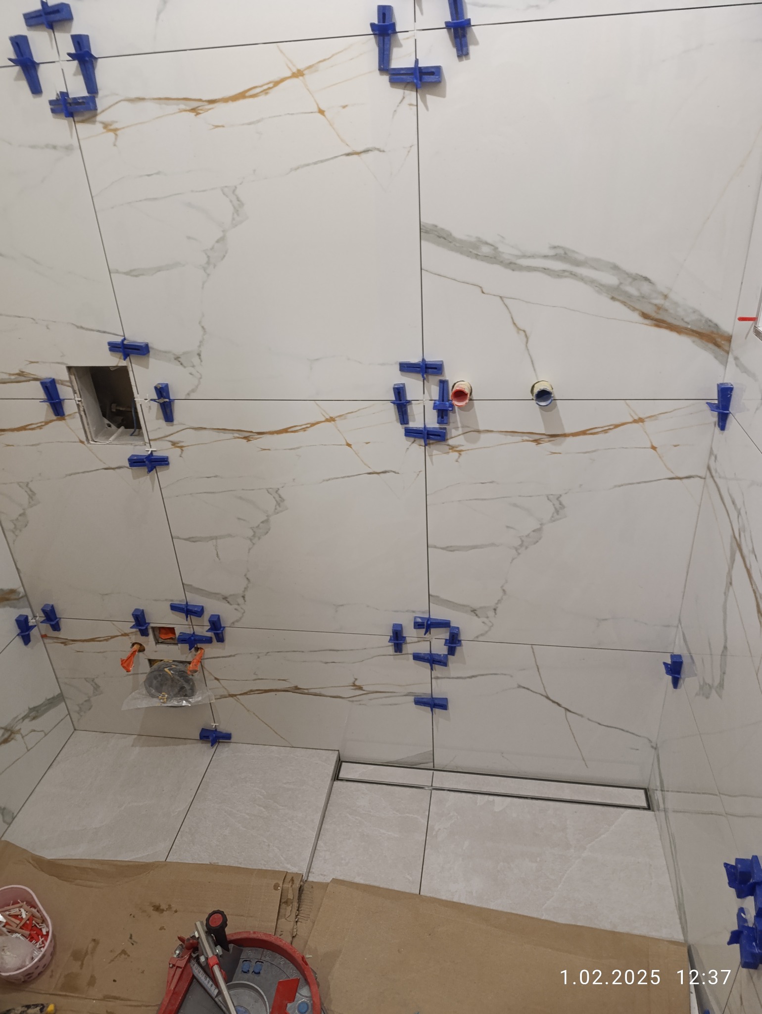 Układanie płytek imitujących marmur w łazience, z użyciem klipsów poziomujących. Widoczny odpływ liniowy i narzędzia glazurnicze.