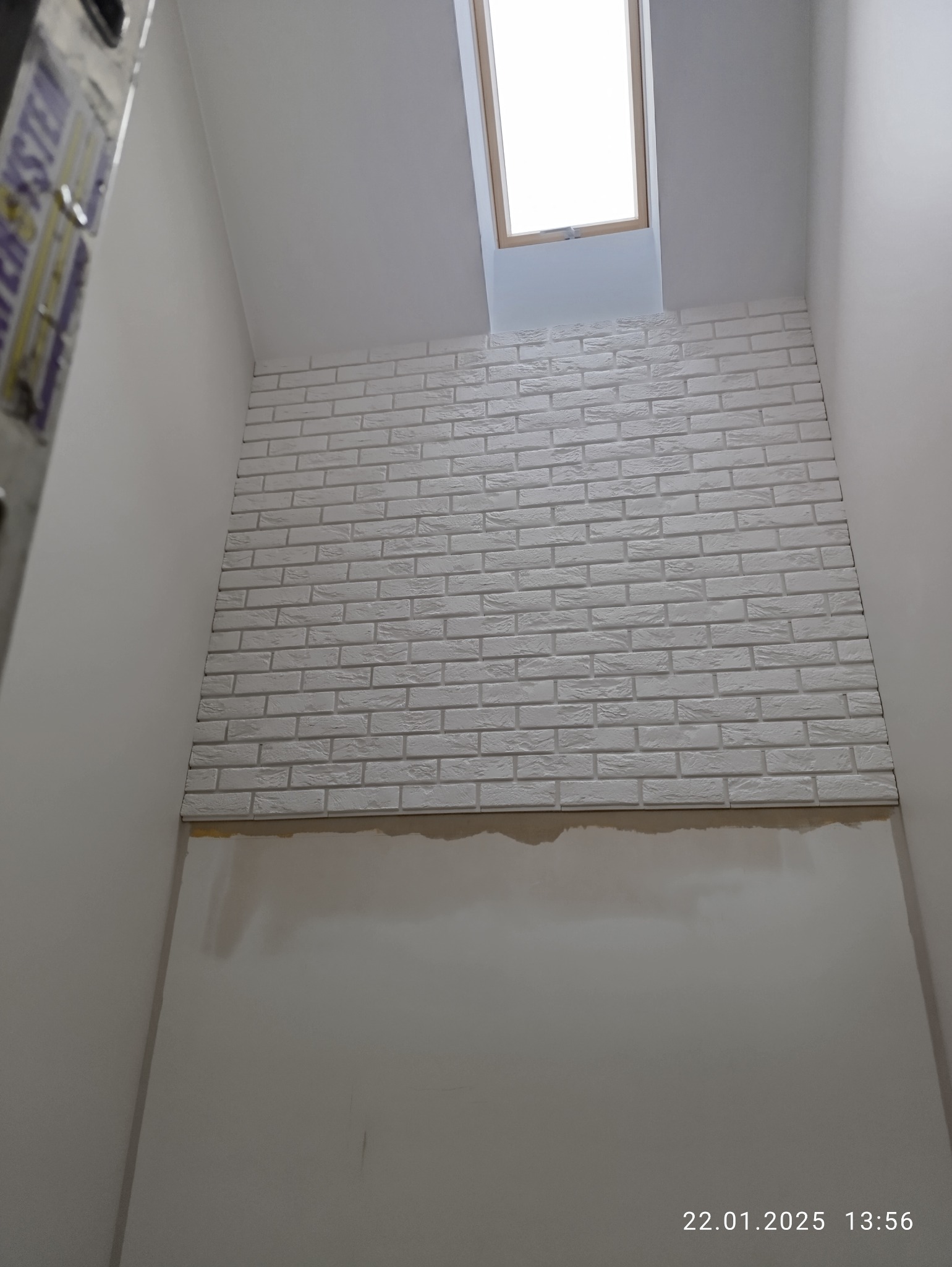 Montaż białej cegły dekoracyjnej na ścianie pod oknem dachowym. Widoczny fragment drabiny. Prace wykończeniowe w jasnym pomieszczeniu.