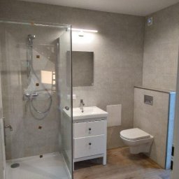 KOREK TEAM PROJEKT SPÓŁKA Z OGRANICZONĄ ODPOWIEDZIALNOŚCIĄ - Jasna łazienka po remoncie: kabina prysznicowa ze szklanymi drzwiami, biała szafka z umywalką, wisząca toaleta, szare płytki na ścianach i drewnopodobna podłoga.