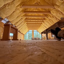 KOREK TEAM PROJEKT SPÓŁKA Z OGRANICZONĄ ODPOWIEDZIALNOŚCIĄ - Ocieplone poddasze domu szkieletowego z widoczną konstrukcją drewnianą, izolacją z wełny mineralnej i oknem na szczycie. Widok z poziomu podłogi, fragment butów w kadrze.