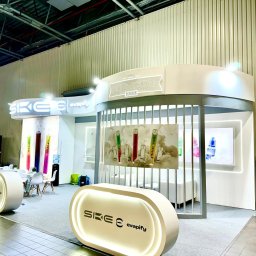 KOREK TEAM PROJEKT SPÓŁKA Z OGRANICZONĄ ODPOWIEDZIALNOŚCIĄ - Jasny, nowoczesny pawilon wystawienniczy SKE Crystal Bar z podświetlanymi elementami, stołami i krzesłami, prezentujący produkty w formie plakatów i ekspozycji.