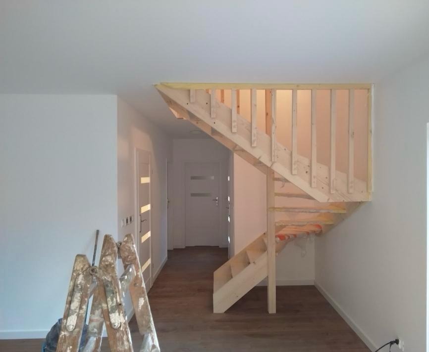 Wnętrze domu w trakcie budowy: surowe drewniane schody z barierkami, drabina malarska, białe ściany i drzwi. Podłoga z paneli. Widok na korytarz.