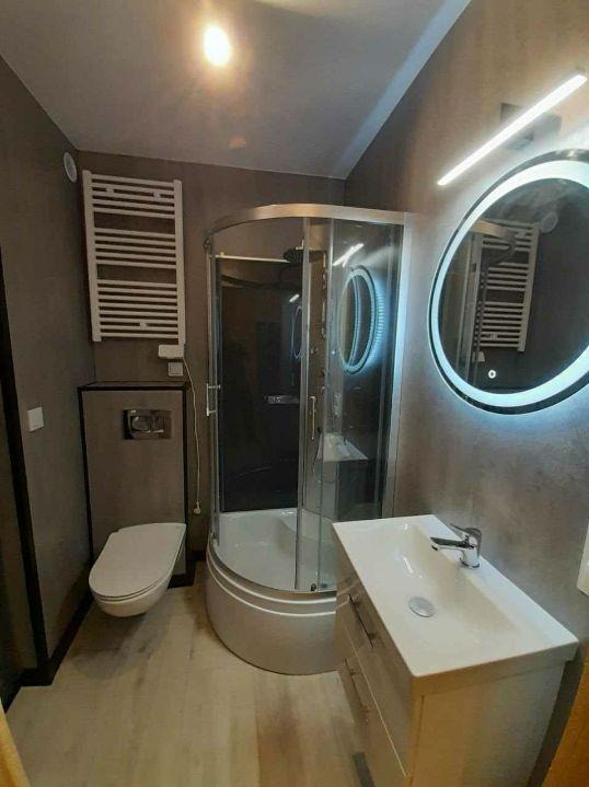 Nowoczesna łazienka w szarościach: kabina prysznicowa z czarnym szkłem, podświetlane lustro, wisząca toaleta i grzejnik drabinkowy. Minimalistyczny design.