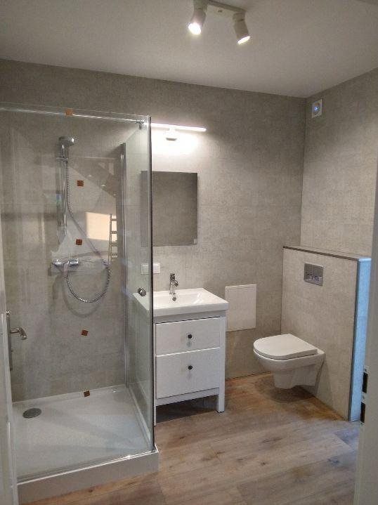 Jasna łazienka po remoncie: kabina prysznicowa ze szklanymi drzwiami, biała szafka z umywalką, wisząca toaleta, szare płytki na ścianach i drewnopodobna podłoga.