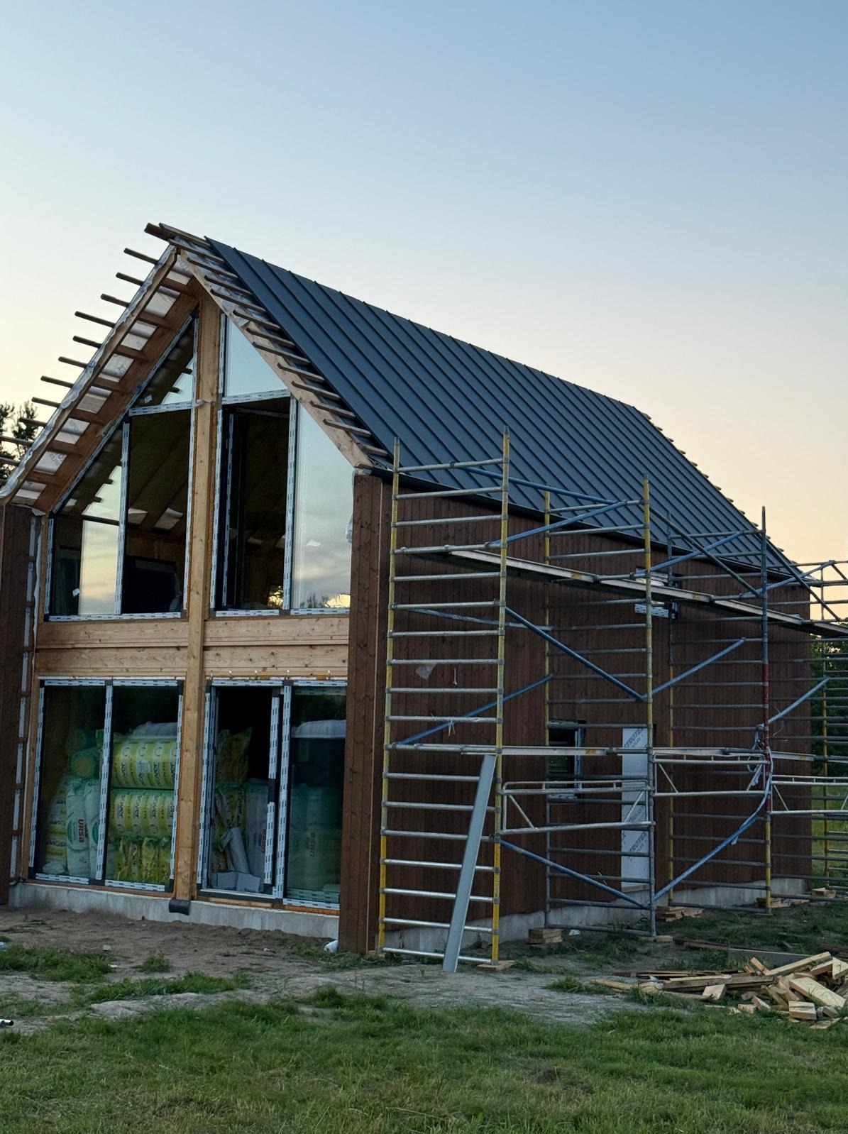 Wznoszenie domu szkieletowego: brązowa elewacja, duże okna z izolacją, metalowy dach i rusztowania w tle, uchwycone o zmierzchu.