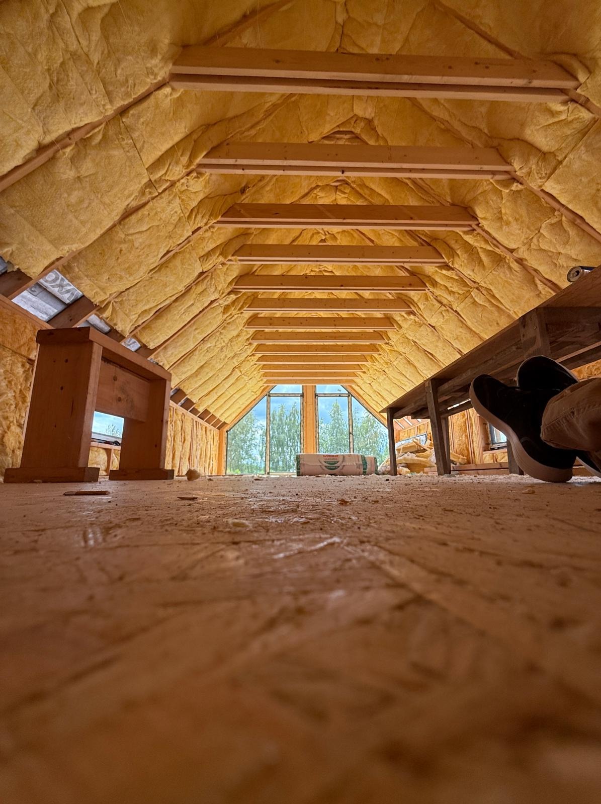 Ocieplone poddasze domu szkieletowego z widoczną konstrukcją drewnianą, izolacją z wełny mineralnej i oknem na szczycie. Widok z poziomu podłogi, fragment butów w kadrze.