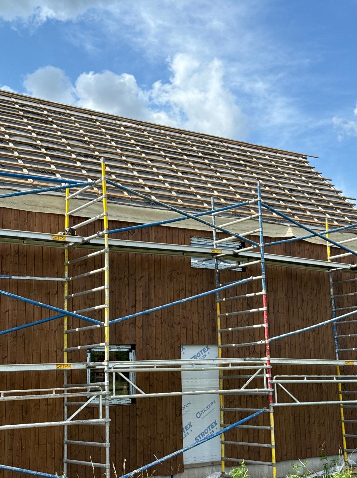 Budowa domu szkieletowego: drewniana elewacja z oknem, rusztowanie i częściowo ułożone deski na dachu pod błękitnym niebem. Widoczny etap prac.
