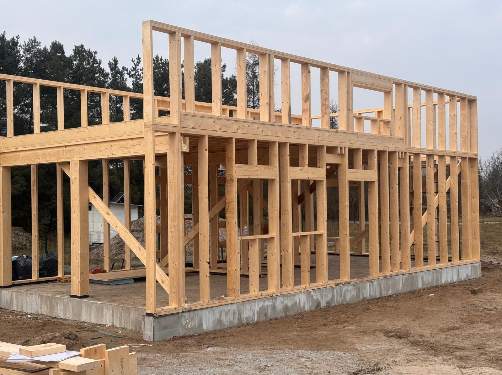 Surowa konstrukcja domu szkieletowego z jasnego drewna na betonowym fundamencie, widok z zewnątrz, w tle drzewa i częściowo widoczny budynek.