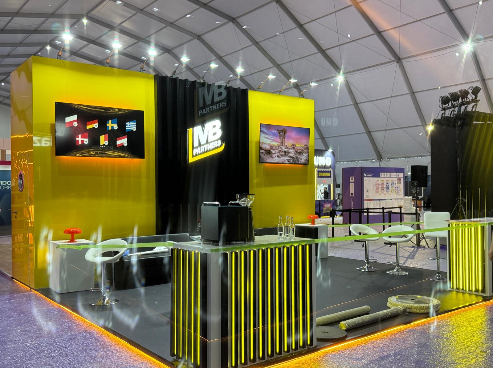 Żółto-czarne stoisko targowe z logo firmy MB Partners, oświetleniem LED i ekranami prezentującymi flagi oraz krajobraz. Nowoczesny design, jasne oświetlenie.
