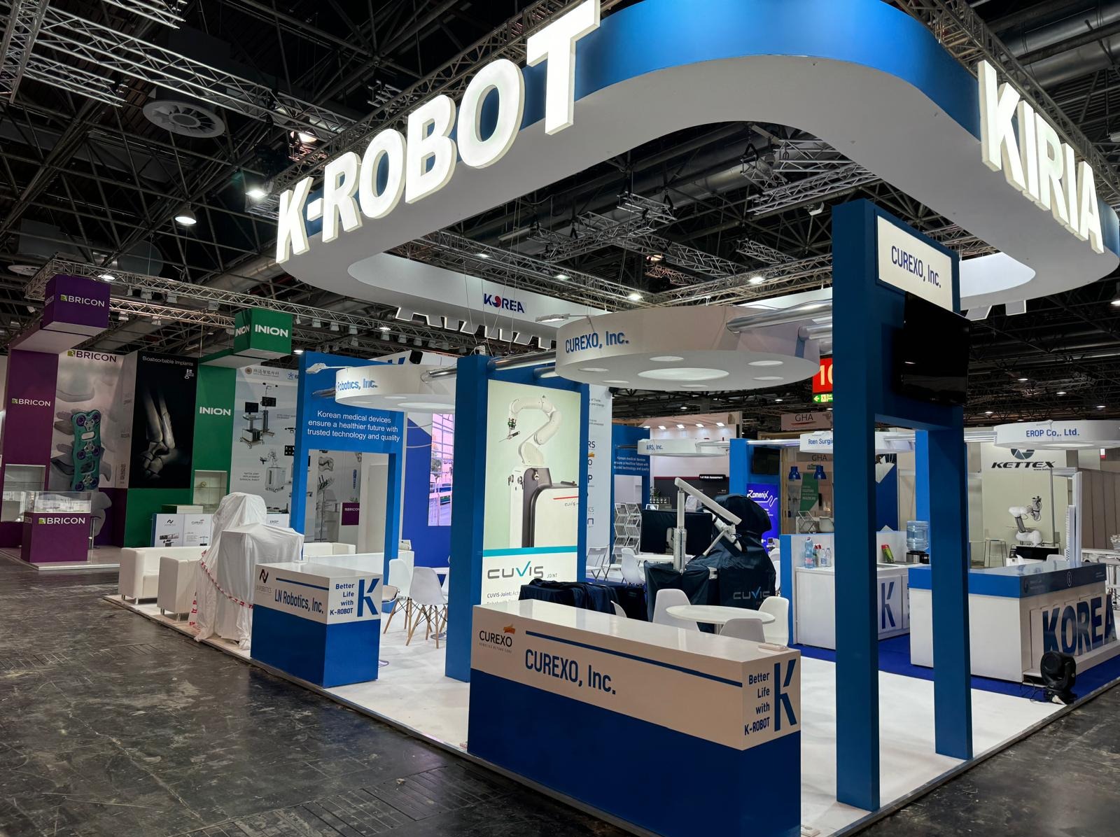 Stoisko targowe K-ROBOT z logo Curexo, Inc. Prezentacja robotów medycznych i urządzeń chirurgicznych. Biało-niebieska kolorystyka, oświetlenie LED, nowoczesny design.