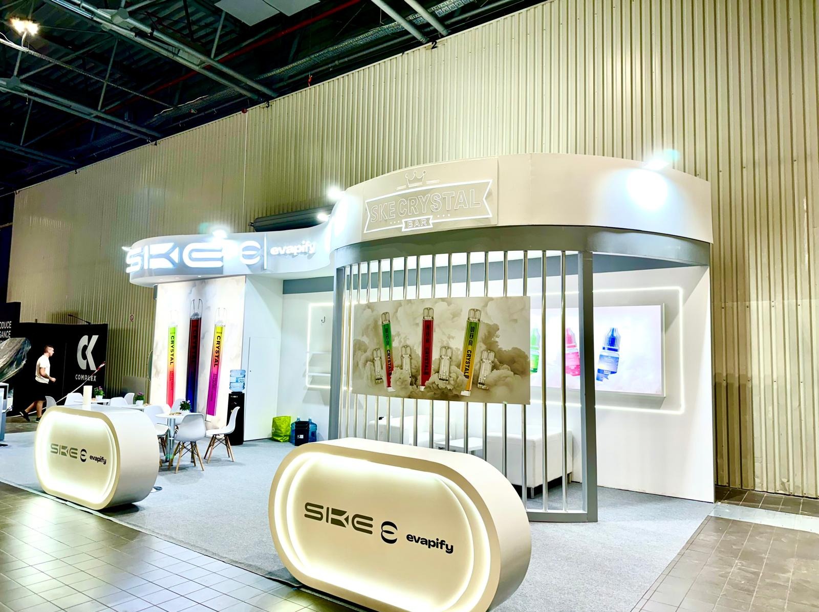 Jasny, nowoczesny pawilon wystawienniczy SKE Crystal Bar z podświetlanymi elementami, stołami i krzesłami, prezentujący produkty w formie plakatów i ekspozycji.