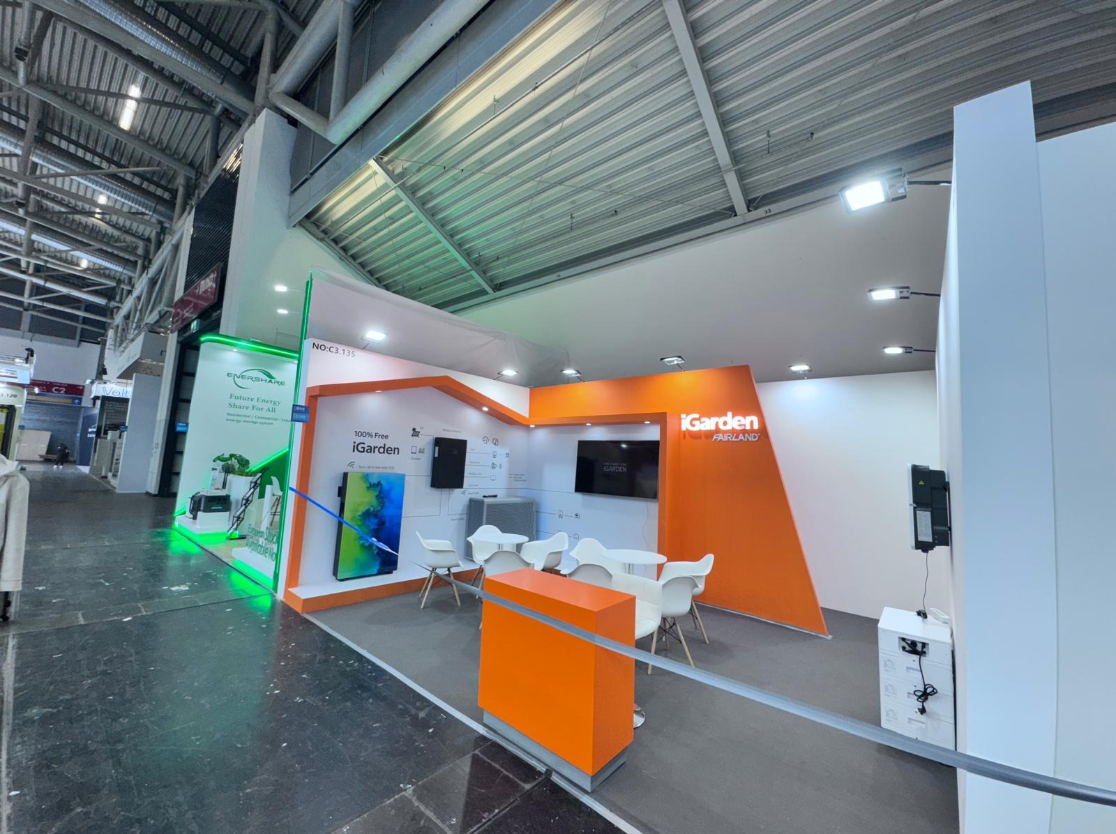 Nowoczesne stoisko targowe iGarden Fairland z pomarańczowymi akcentami, białymi meblami i ekranem. Widok z perspektywy zwiedzającego, oświetlenie sufitowe.