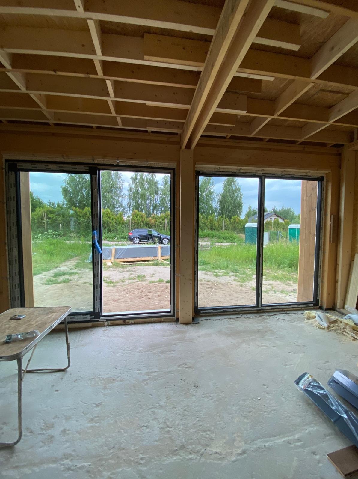 Wnętrze domu szkieletowego w trakcie budowy. Widoczne drewniane elementy konstrukcyjne, duże okna przesuwne z widokiem na zewnątrz i surowa betonowa podłoga.