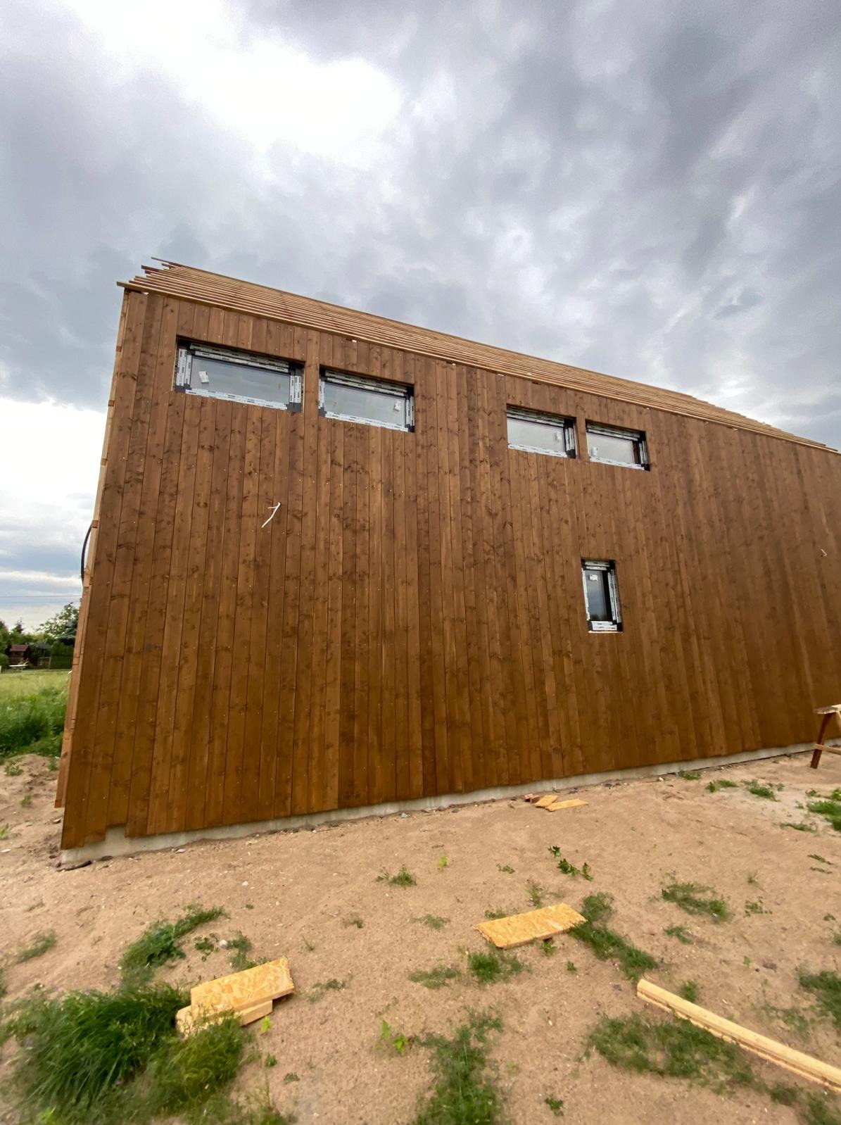 Nowoczesny dom szkieletowy z brązową elewacją z desek, widoczne okna z PCV i fragment dachu, na zewnątrz teren budowy z resztkami materiałów.