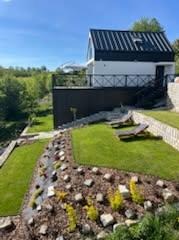 Nowoczesny dom z czarnym dachem i balkonem, otoczony zielonym trawnikiem z kamieniami. Ujęcie z dołu podkreśla architekturę budynku i zagospodarowanie terenu.