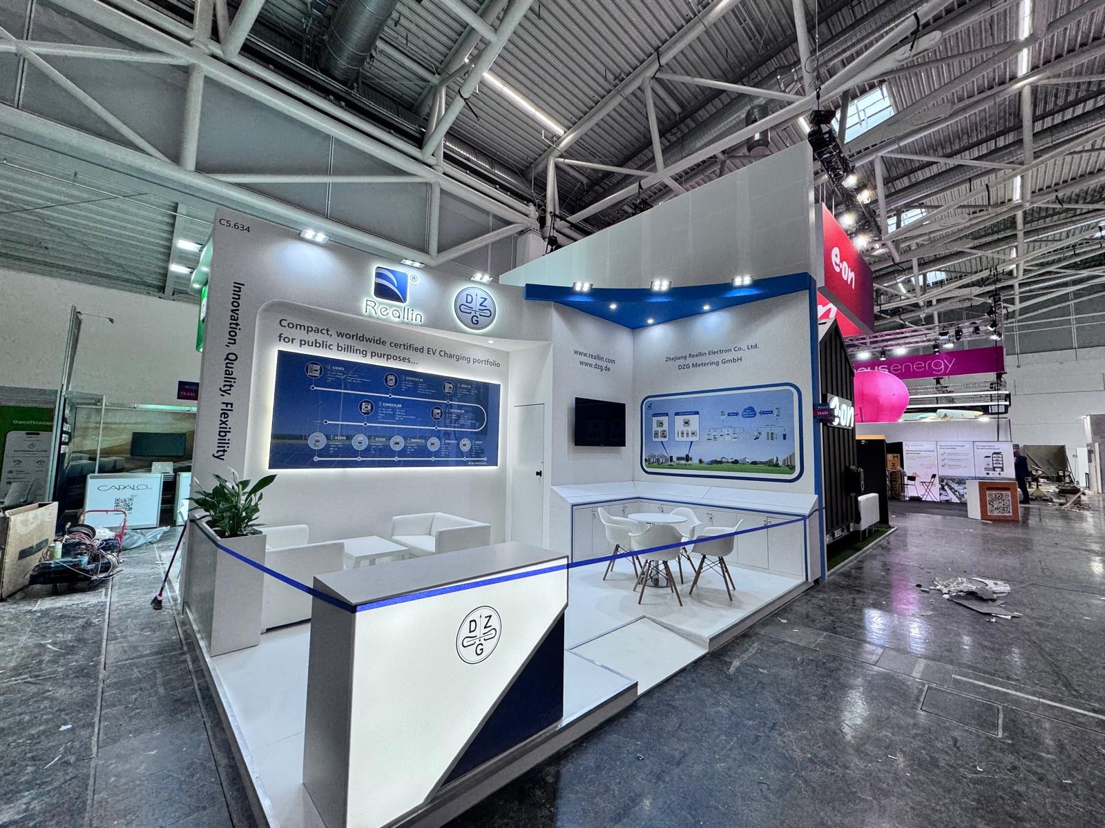 Stoisko targowe Reallin z logo DZG, prezentujące rozwiązania EV. Nowoczesny design, białe meble, grafiki informacyjne. Widok na halę targową z oświetleniem.
