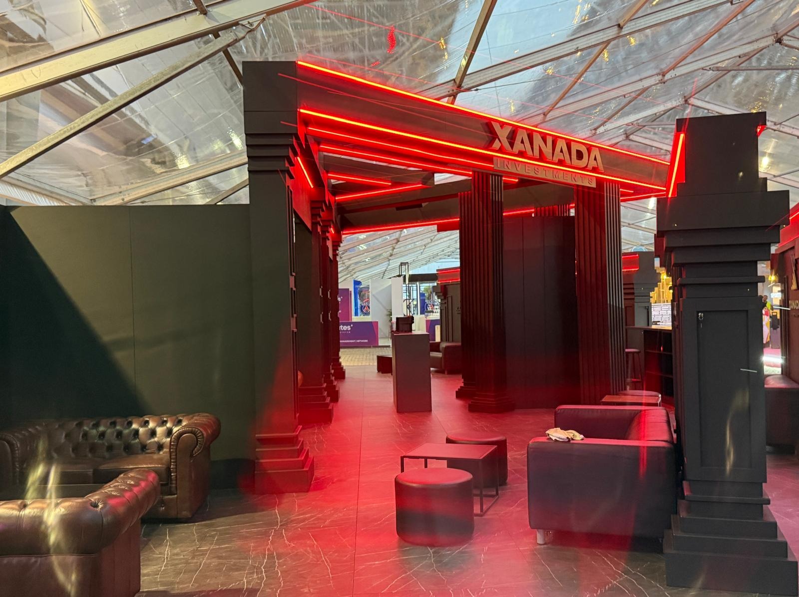 Nowoczesne stoisko targowe Xanada Investments z czerwonym neonem, czarnymi kolumnami i skórzanymi sofami, oświetlone czerwonym światłem, pod przezroczystym namiotem.