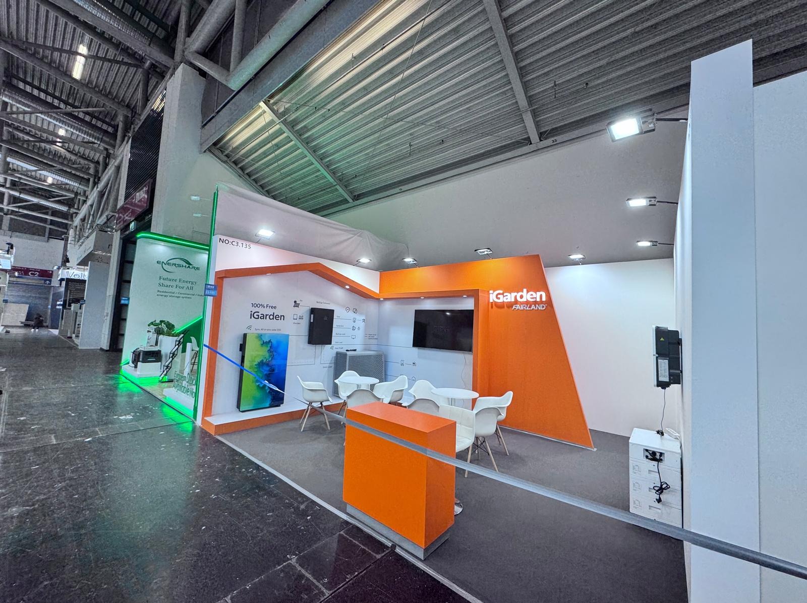 Stoisko targowe iGarden Fairland z ekranami, białymi krzesłami i pomarańczowymi elementami, oświetlone światłem LED, widoczne z perspektywy lekko z dołu.