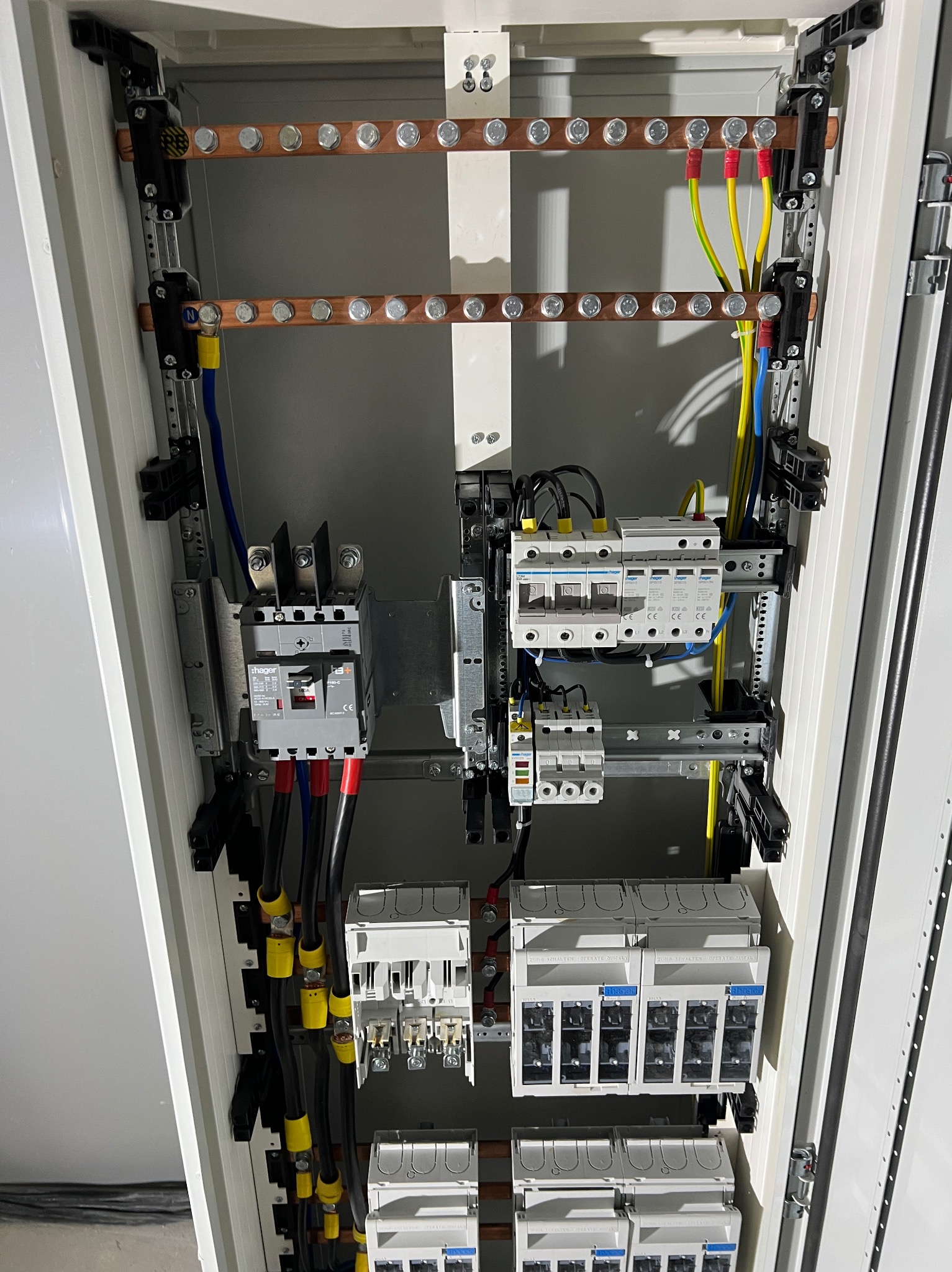 Rozdzielnica elektryczna w metalowej obudowie, widoczne bezpieczniki, przewody w kolorach: żółtym, niebieskim i czarnym. Elementy instalacji zamontowane wewnątrz szafy.