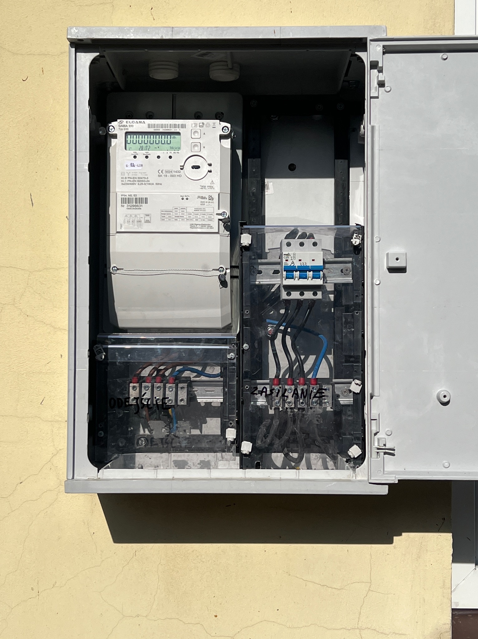 Otwarta skrzynka elektryczna z licznikiem i bezpiecznikami na elewacji budynku. Widoczne przewody i elementy instalacji. Jasne światło dzienne.