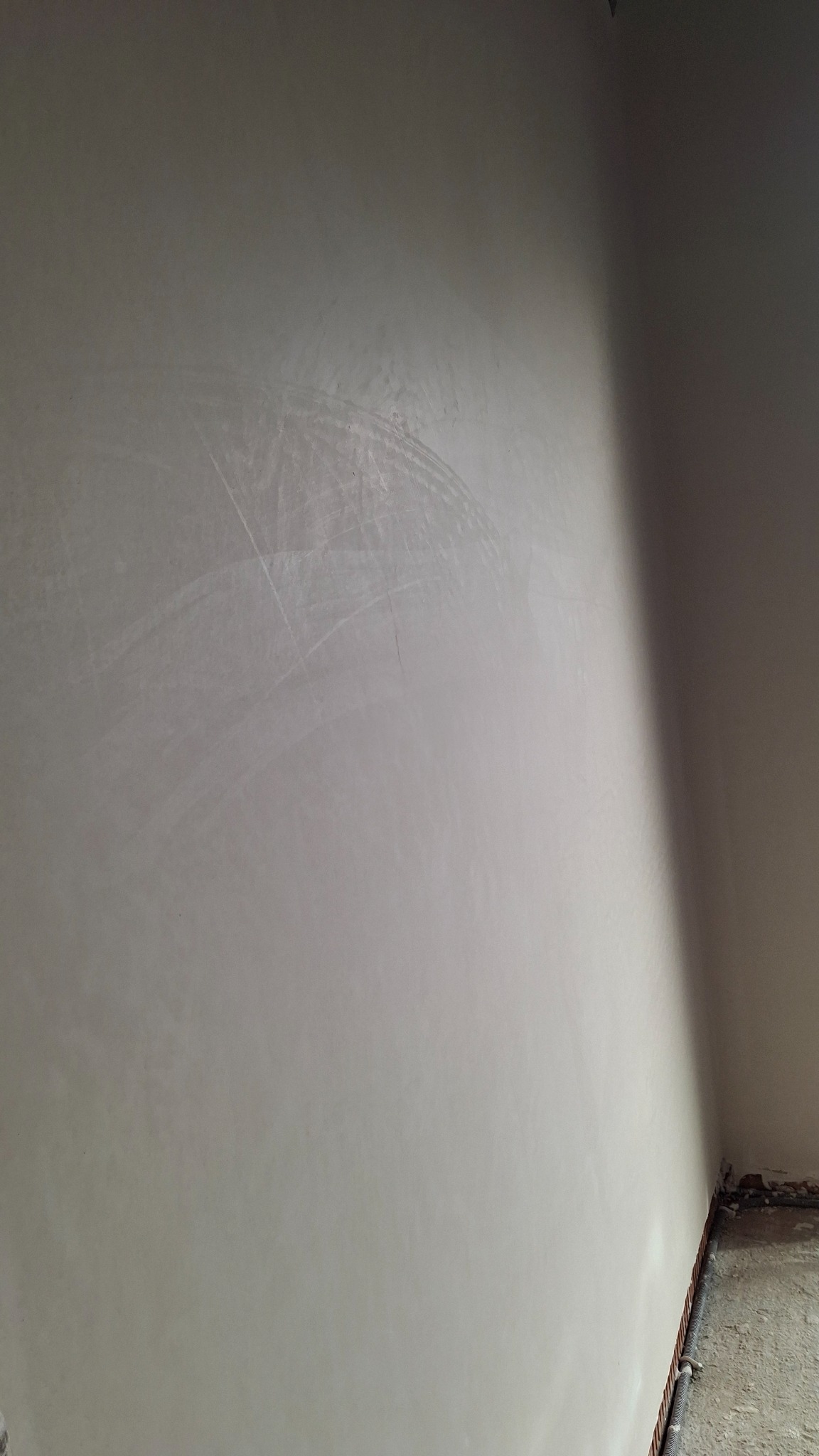 Gładka, jasna ściana po nałożeniu tynku maszynowego, widoczne delikatne ślady po szpachlowaniu. Fragment podłogi z rurami w rogu pomieszczenia.