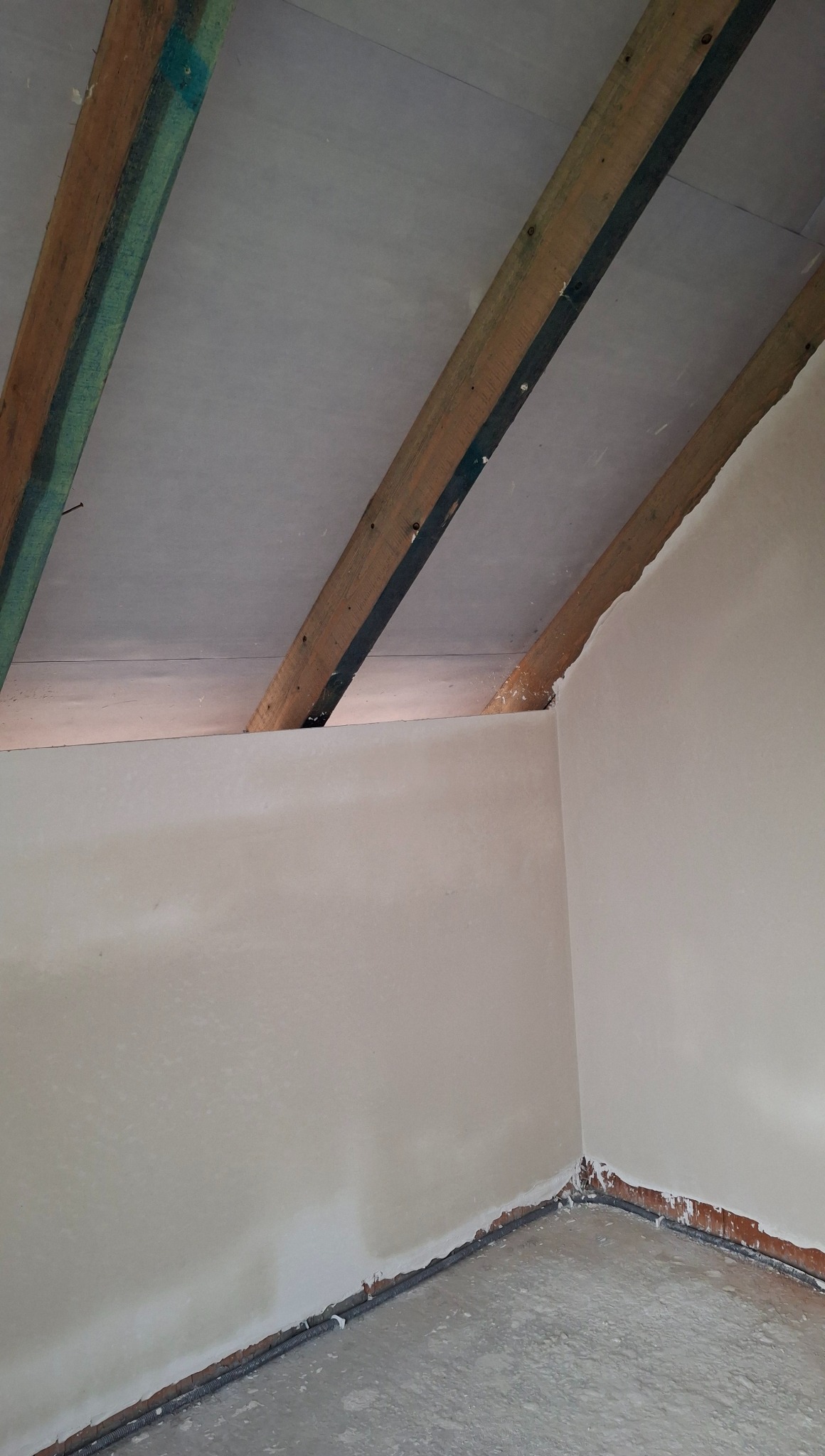 Świeżo otynkowane poddasze z drewnianą konstrukcją dachu. Widoczne niedoskonałości przy podłodze i suficie. Szare tynki na ścianach i suficie.