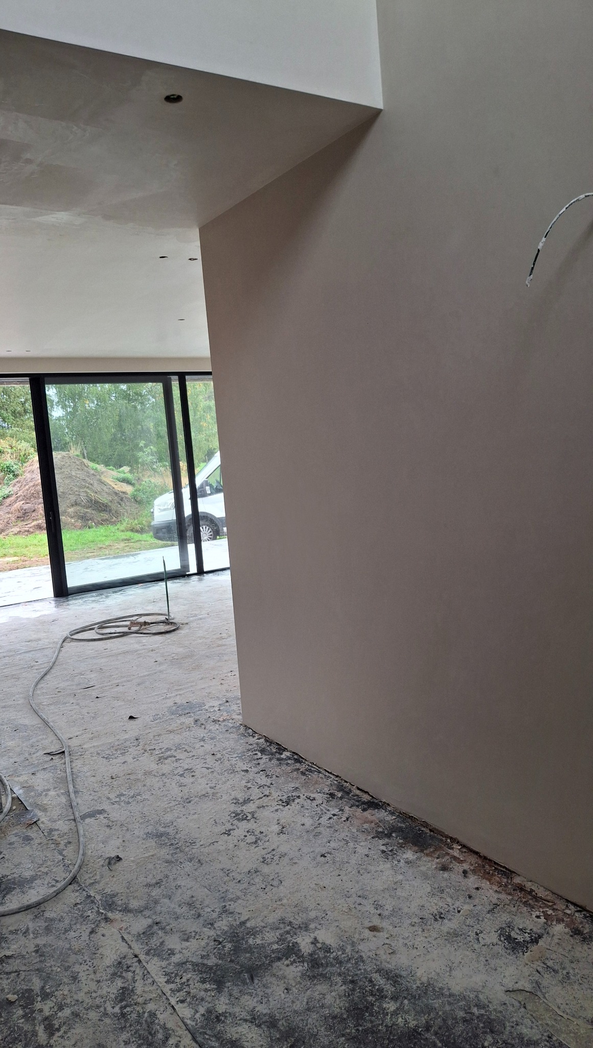 Gładka, beżowa ściana po tynkowaniu maszynowym w surowym wnętrzu z dużym oknem i widocznym wężem instalacyjnym na podłodze. Na zewnątrz zaparkowany biały bus.