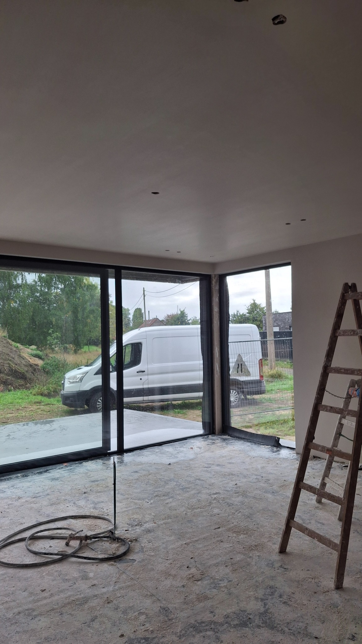 Wnętrze po tynkowaniu maszynowym: gładkie ściany, duże okna z widokiem na zewnątrz, drabina oparta o ścianę, przewody na podłodze. Widoczne ślady po pracach.