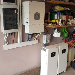 Kacper Dymitrasz - Zestaw: rozdzielnica, inwerter i dwa magazyny energii na kółkach, zamontowane na ścianie w garażu. Widoczne przewody i rury instalacyjne. Półka z narzędziami w tle.