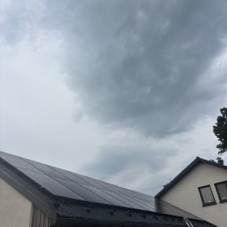 Kacper Dymitrasz - Panele słoneczne na dachu domu jednorodzinnego w pochmurny dzień. Widoczna drabina oparta o dach. Ujęcie z dołu, podkreślające instalację.