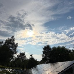 Kacper Dymitrasz - Błyszczące panele słoneczne odbijają niebo z chmurami, otoczone zielenią drzew i krzewów. Instalacja wkomponowana w krajobraz, widok z dołu.