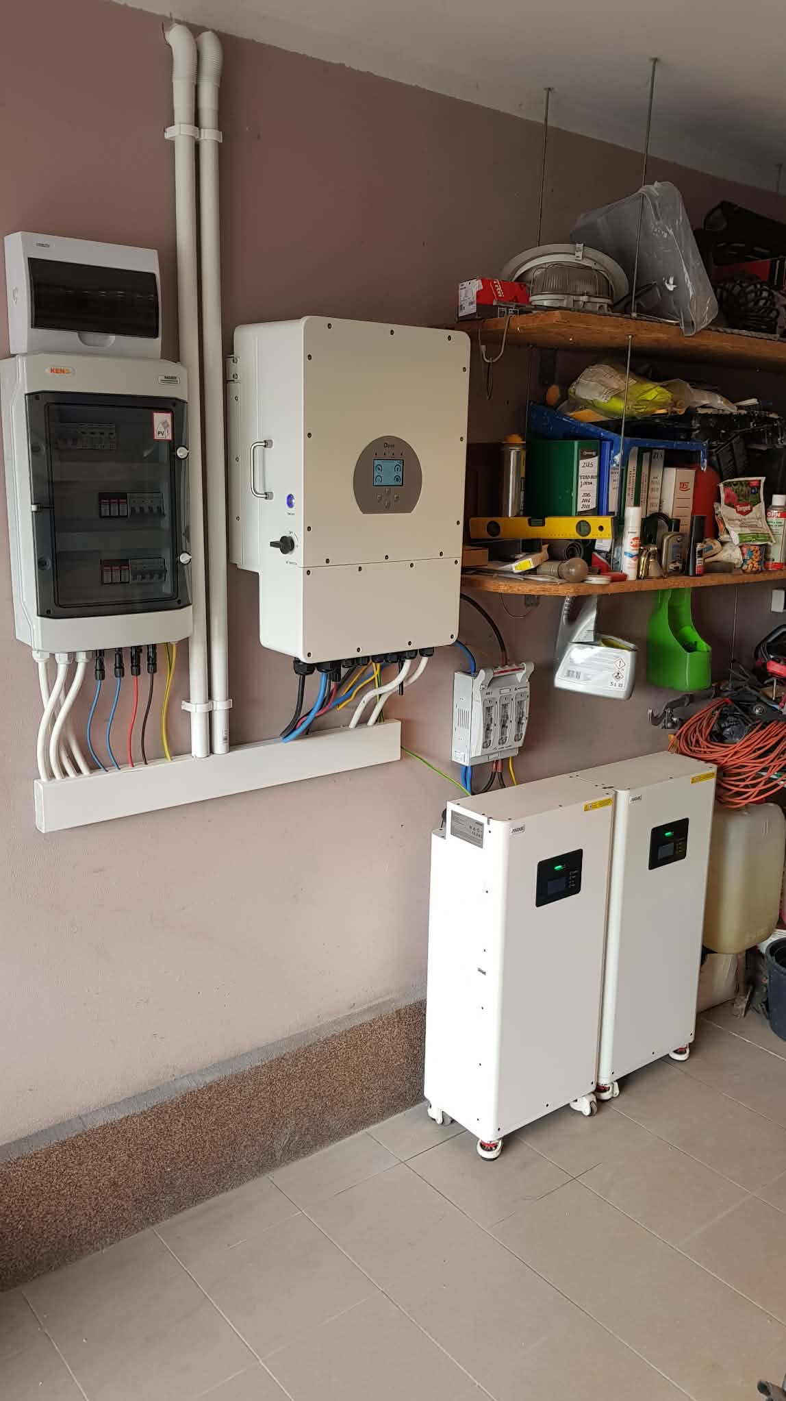 Zestaw: rozdzielnica, inwerter i dwa magazyny energii na kółkach, zamontowane na ścianie w garażu. Widoczne przewody i rury instalacyjne. Półka z narzędziami w tle.