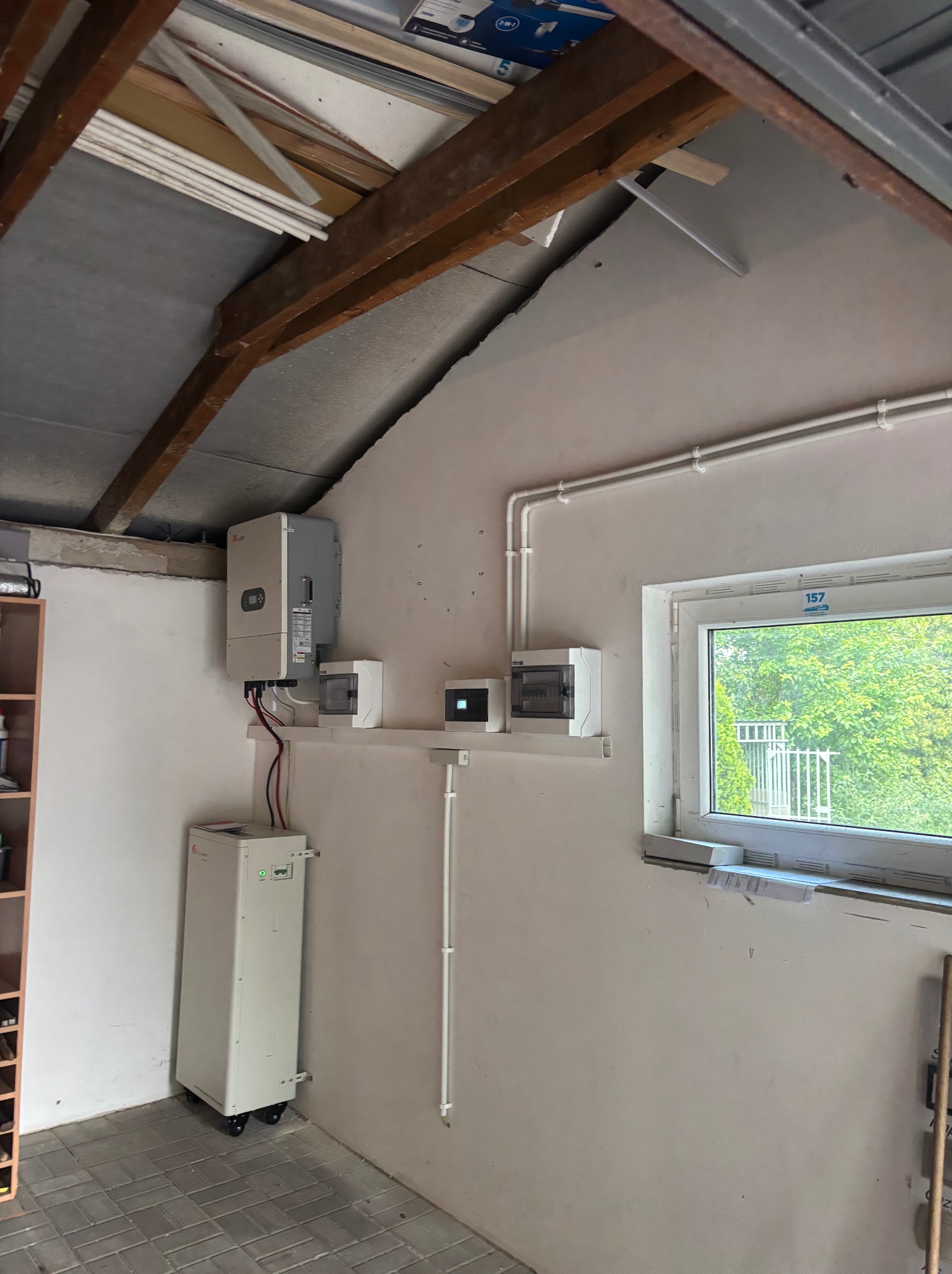 Zamontowany inwerter i magazyn energii w garażu, widoczne okablowanie i zabezpieczenia. Instalacja w pomieszczeniu z oknem, na ścianie białe rury instalacyjne.