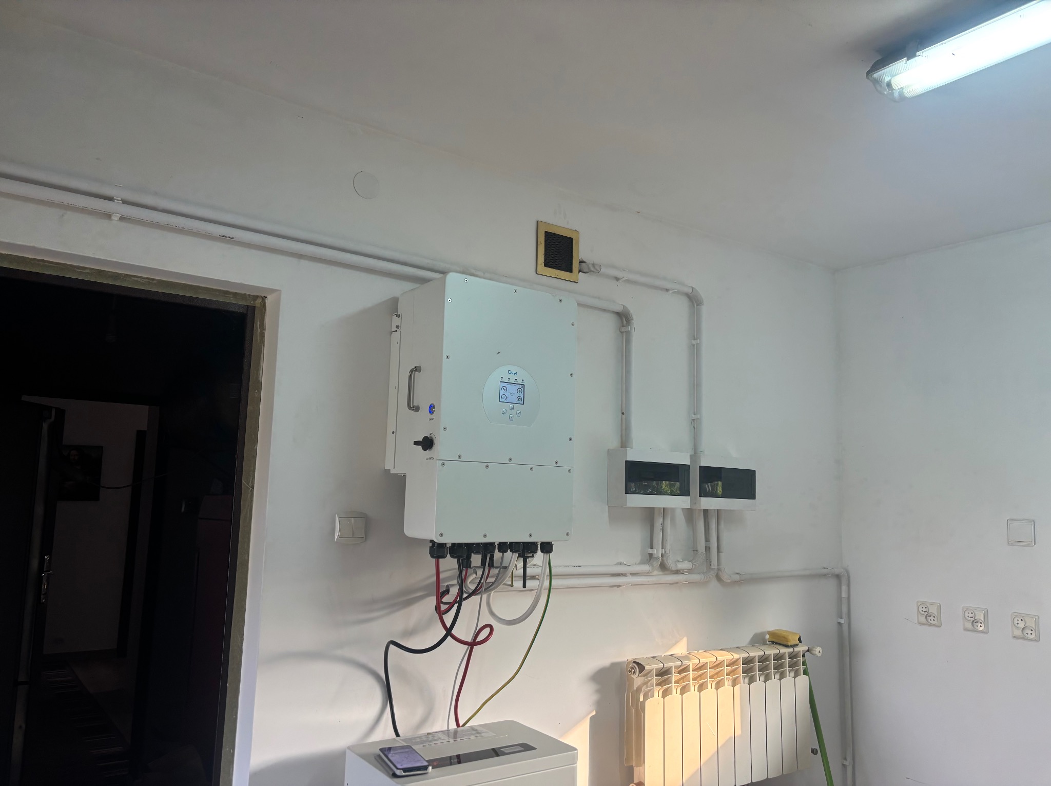 Biały inwerter fotowoltaiczny zamontowany na ścianie wewnątrz pomieszczenia, widoczne przewody, rury instalacyjne i grzejnik. Ujęcie z bliska, wnętrze oświetlone.