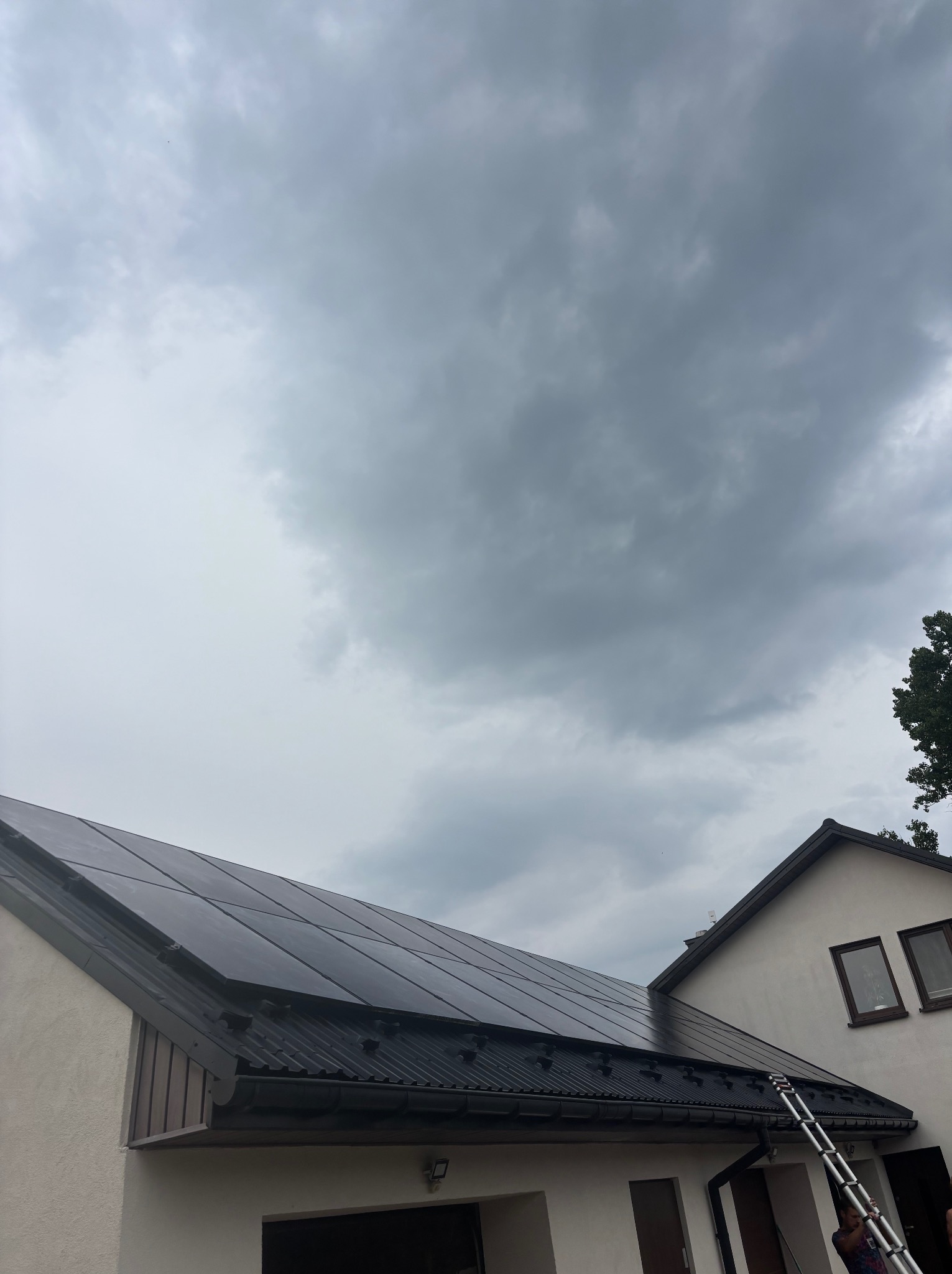 Panele słoneczne na dachu domu jednorodzinnego w pochmurny dzień. Widoczna drabina oparta o dach. Ujęcie z dołu, podkreślające instalację.