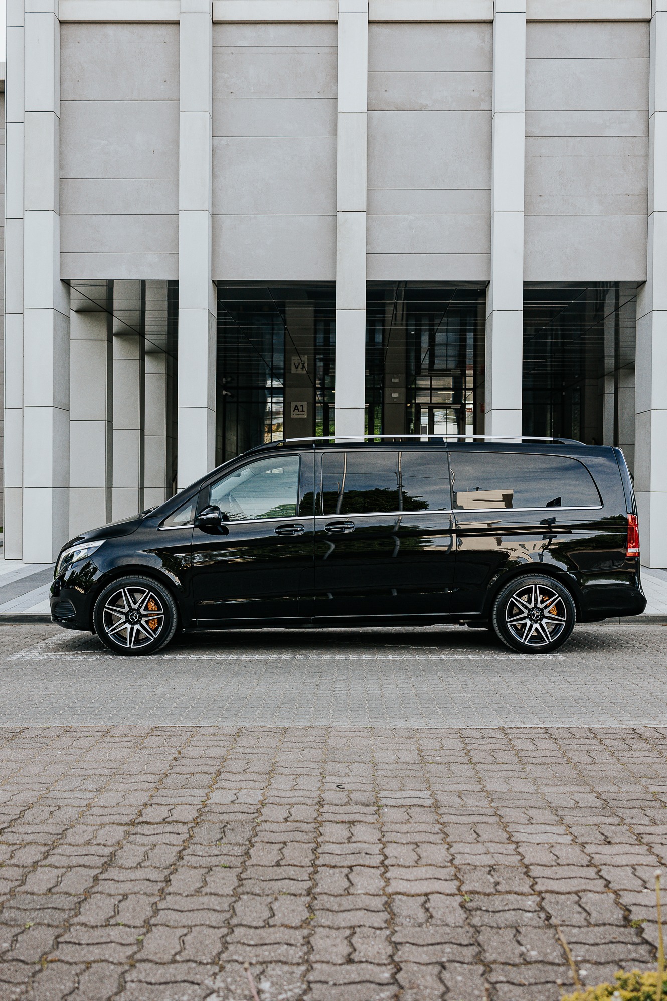 Elegancki, czarny bus Mercedes V-klasy zaparkowany na brukowanej nawierzchni przed nowoczesnym budynkiem z szarymi panelami. Widok z boku, felgi z logo Mercedesa.