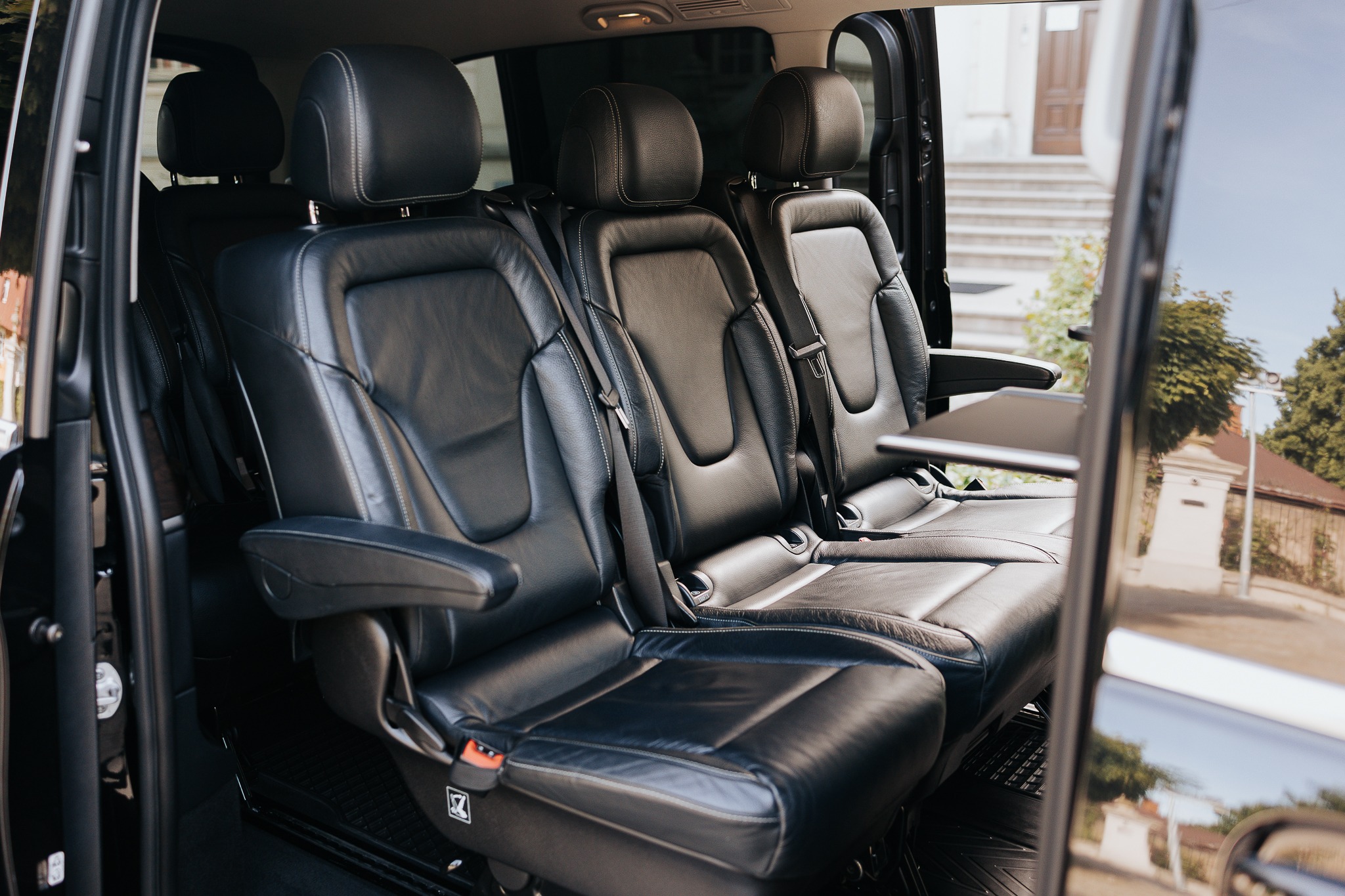 Wnętrze luksusowego busa z czarnymi skórzanymi fotelami, widok z otwartych drzwi na zewnątrz, gdzie widać budynek i zieleń.