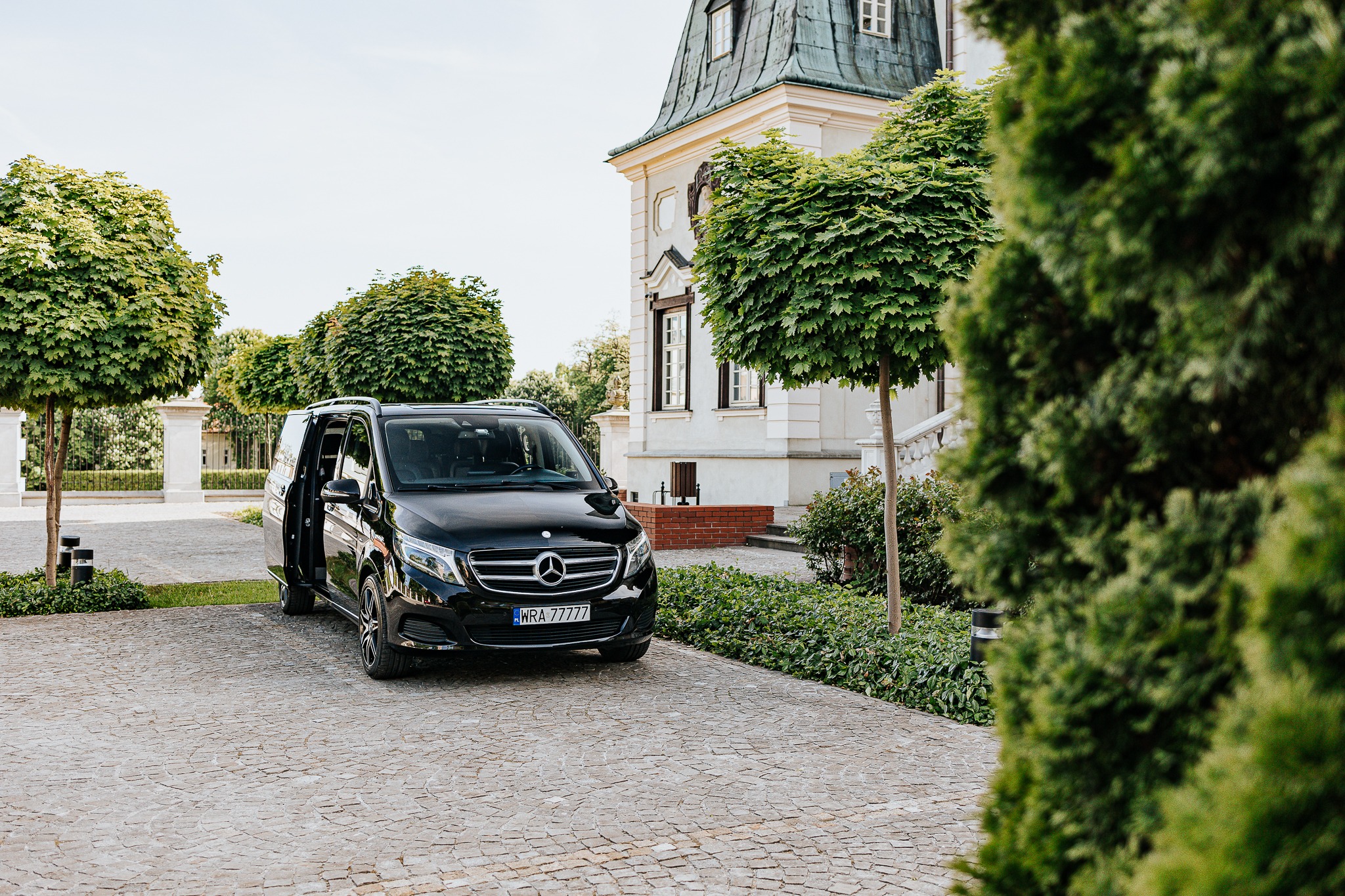 Czarny, luksusowy bus Mercedes V-klasy z otwartymi drzwiami, zaparkowany na brukowanym podjeździe przed eleganckim budynkiem z zielonym dachem i ozdobnymi drzewami.