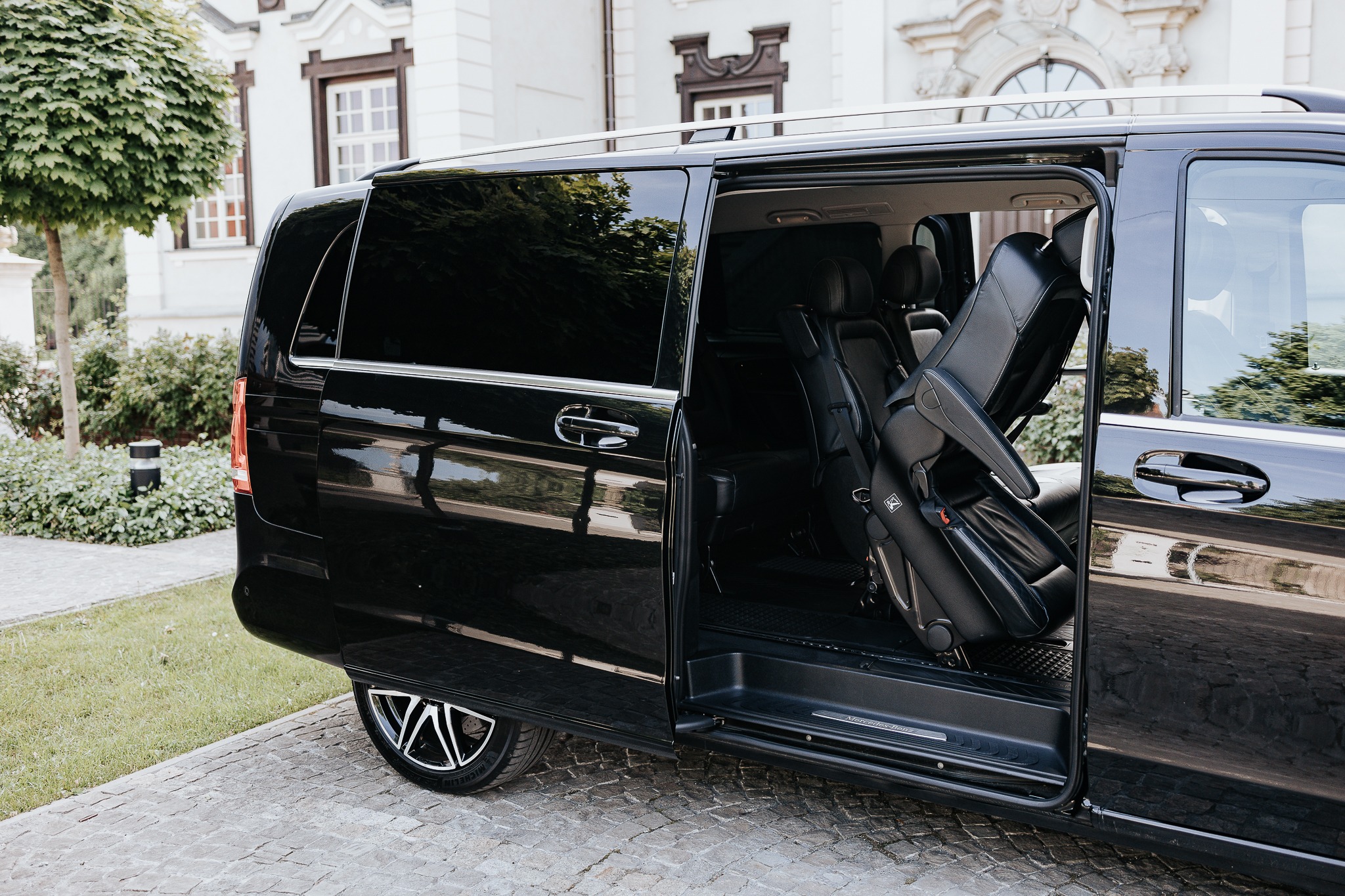 Elegancki, czarny bus z otwartymi drzwiami przesuwnymi, ukazujący luksusowe, skórzane wnętrze. Widoczne detale karoserii i felgi. Tło stanowi budynek i zieleń.