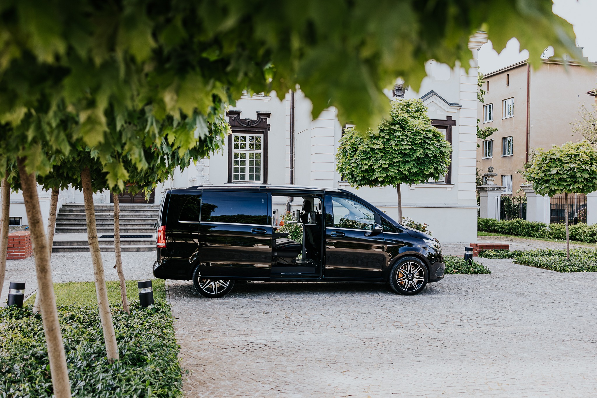 Elegancki, czarny bus Mercedes z otwartymi drzwiami na tle klasycznej architektury i zieleni. Luksusowy transport na specjalne okazje, np. ślub.