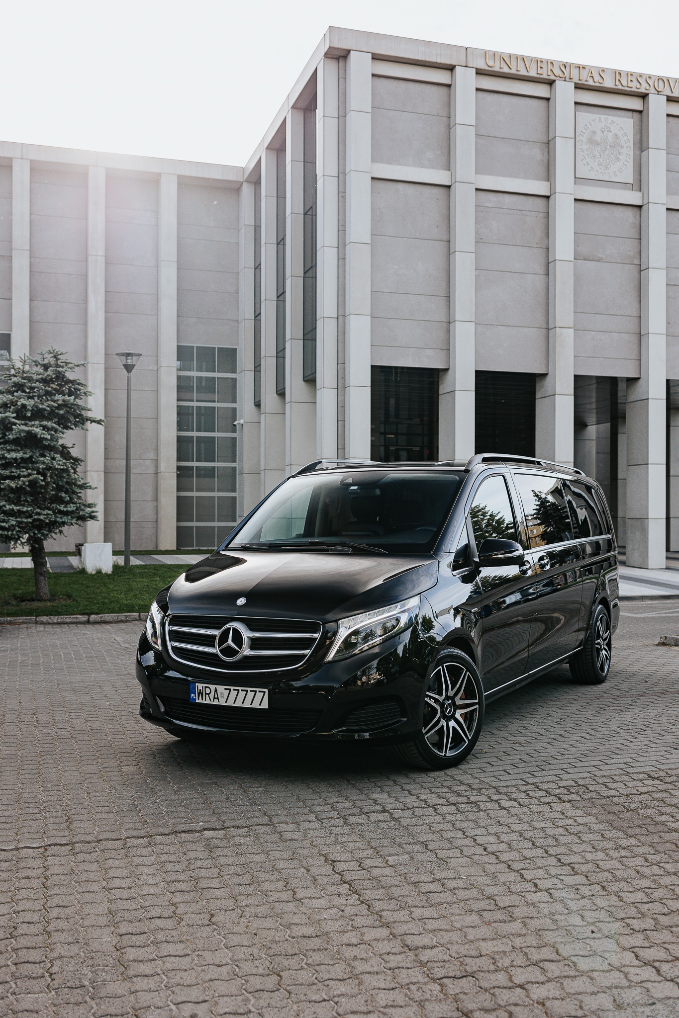 Czarny, luksusowy van Mercedes V-klasy na tle budynku Uniwersytetu Rzeszowskiego. Elegancki pojazd do przewozu osób, z widoczną tablicą rejestracyjną.