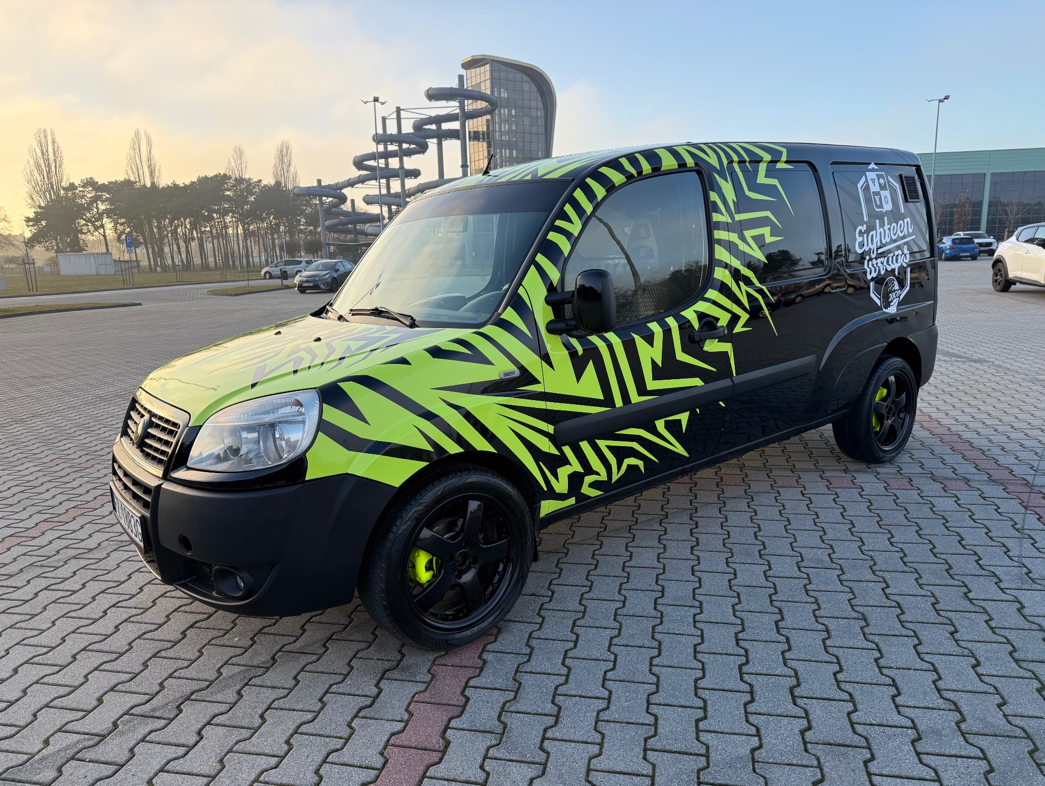 Czarny van z jaskrawozielonym, geometrycznym wzorem oklejenia, zaparkowany na kostce brukowej, z budynkiem aquaparku w tle. Widoczne logo firmy na boku.