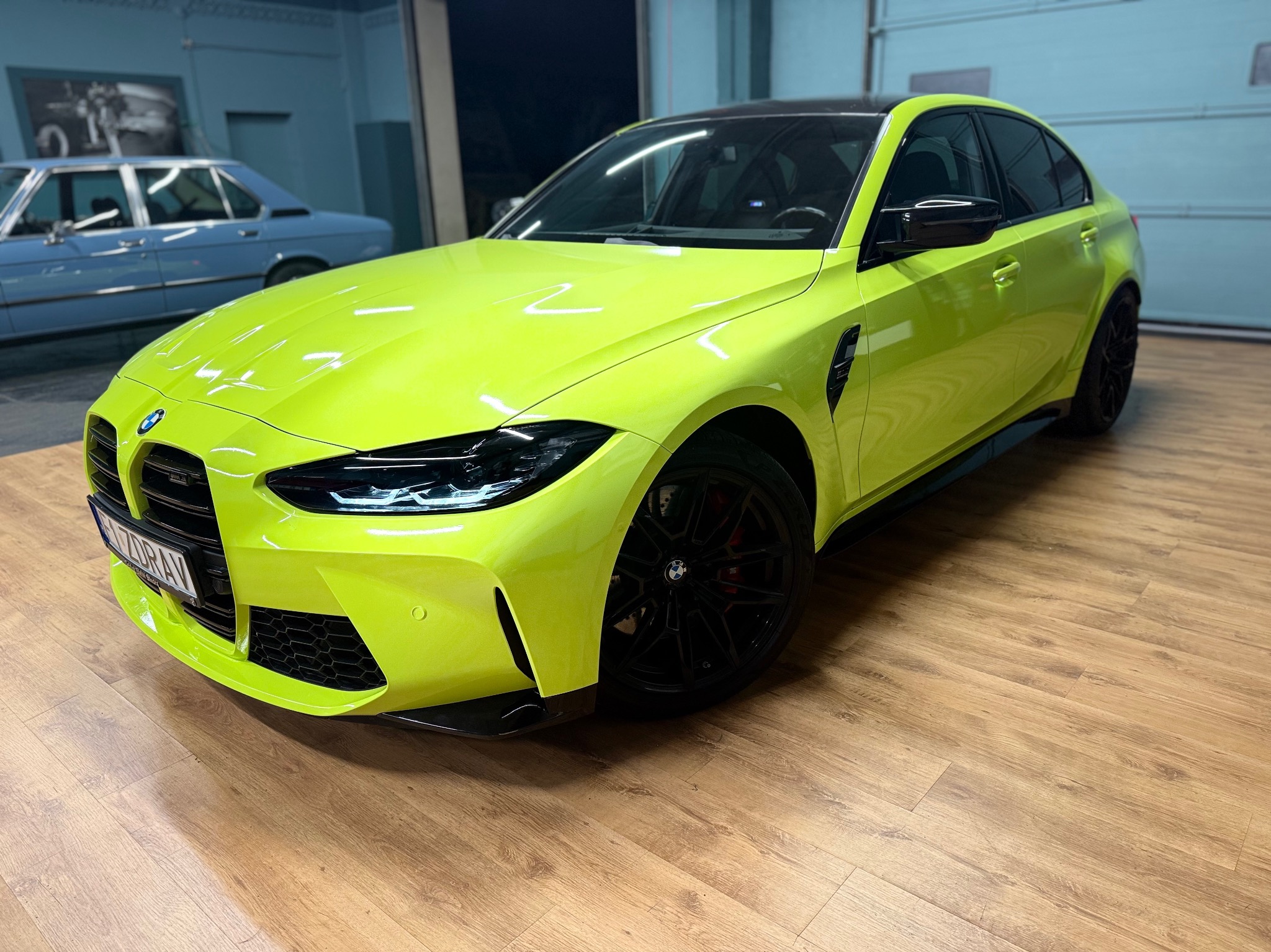 Jaskrawożółte BMW M3 z czarnymi felgami i dachem, zaparkowane w garażu. W tle widoczny klasyczny, niebieski samochód.