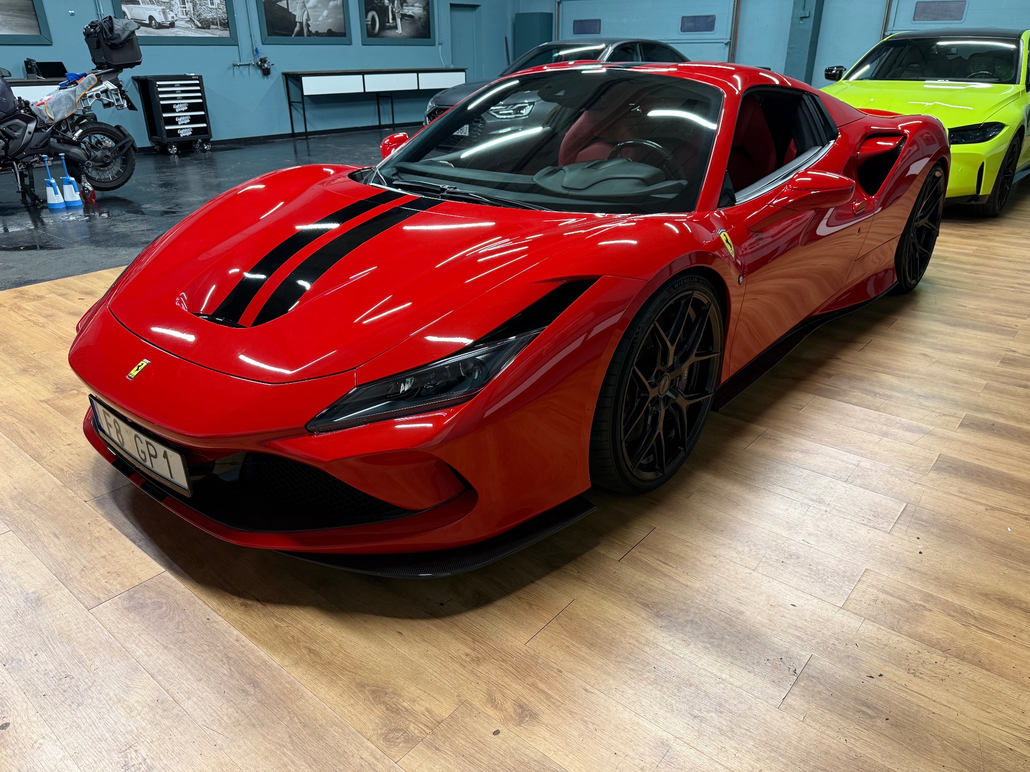 Czerwone Ferrari F8 Tributo z czarnymi pasami na masce, zaparkowane w garażu z innymi autami. Widoczny detal felg i tablicy rejestracyjnej.