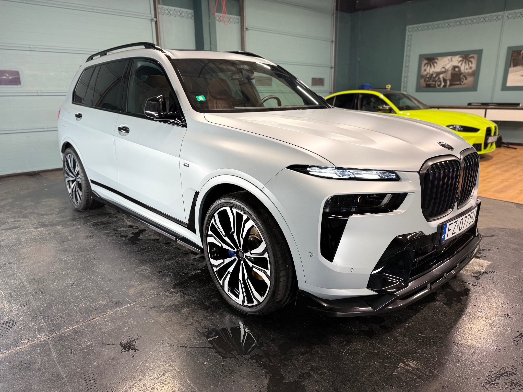 Matowy, biały SUV BMW X7 z czarnymi akcentami i felgami, zaparkowany w garażu. W tle widoczny neonowożółty samochód. Refleksy na podłodze.