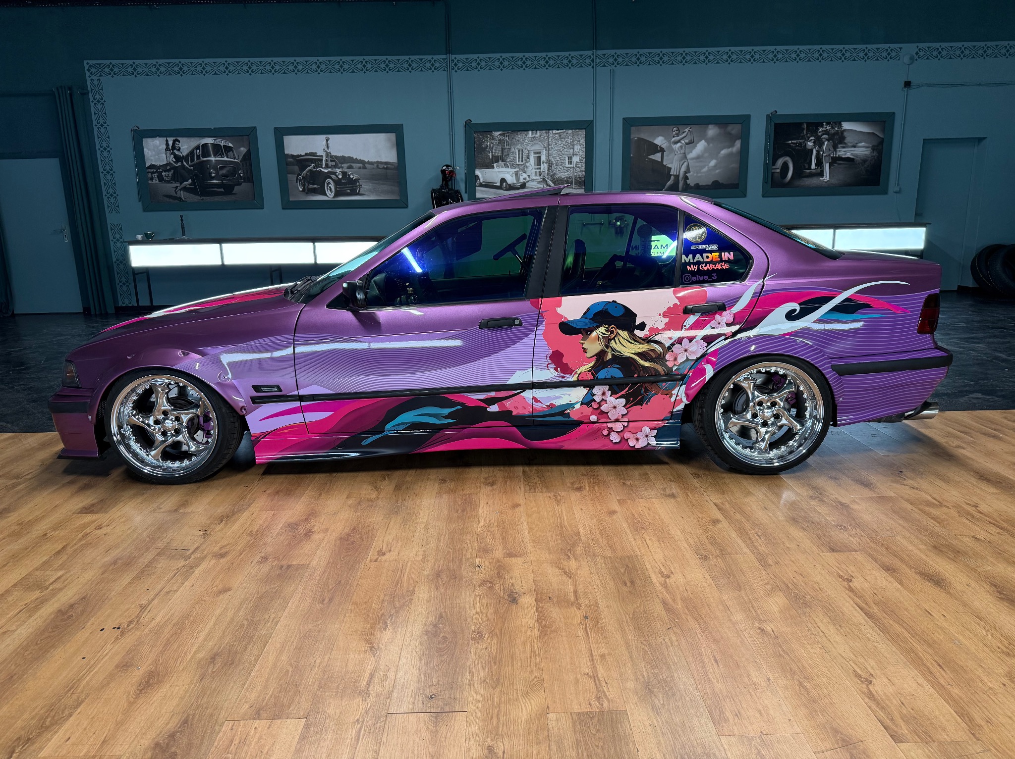 Fioletowy samochód BMW E36 z unikalną grafiką w stylu anime, zaparkowany w warsztacie z drewnianą podłogą i czarno-białymi zdjęciami na ścianie.